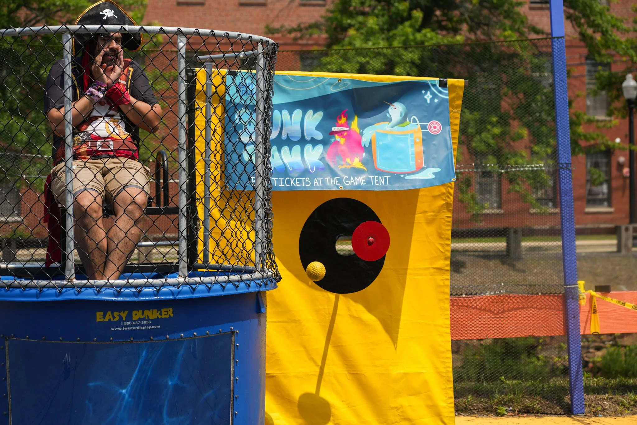 A man dressed as a pirate is inside a dunk tank, sitting on a seat and ready to be dunked. The dunk tank has a blue base and is surrounded by a metal fence. The background features a colorful banner advertising tickets at the game tent, with vibrant