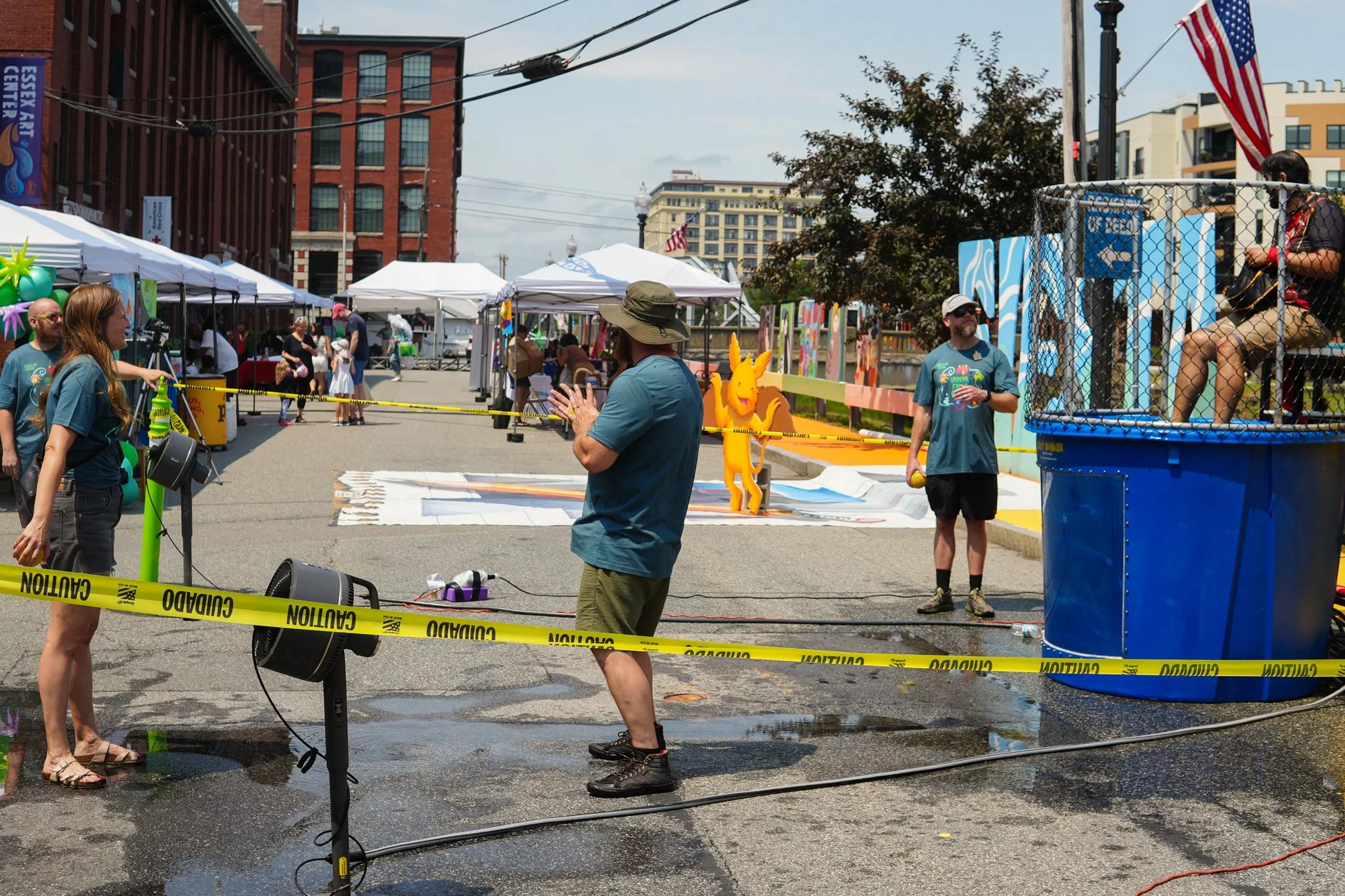 People at an outdoor street fair with tents, colorful artwork, and clown sculptures. Caution tape and wet pavement are visible, indicating ongoing setup or cleanup.