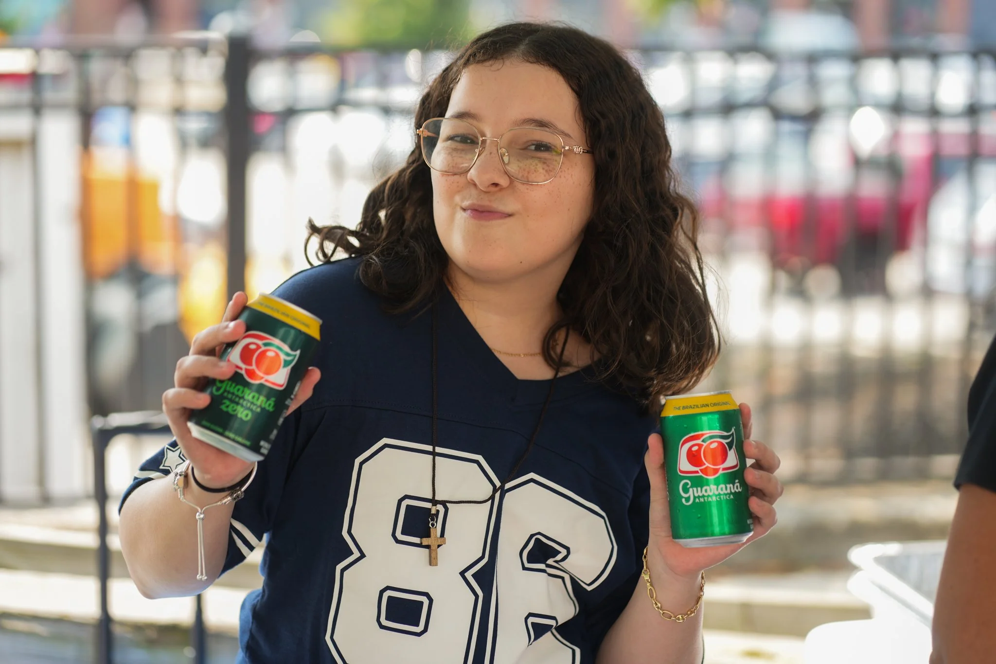 A young woman with glasses and dark curly hair holding two cans of Guaraná Antarctica soda, smiling at the camera.