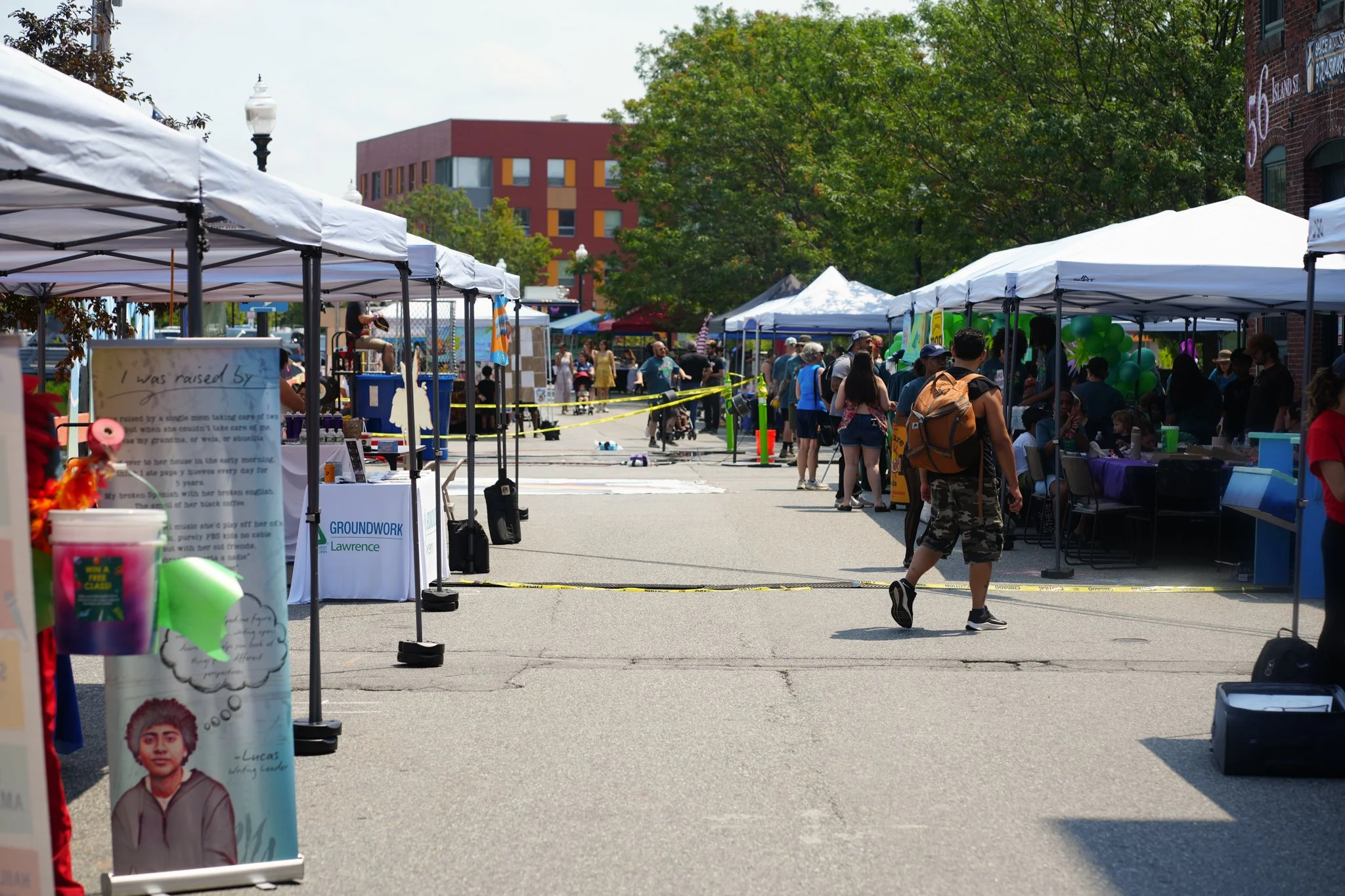 Outdoor street market with multiple vendor tents and people walking and browsing. Tents have signs and decorations, some with balloons. Crowds and trees are in the background.