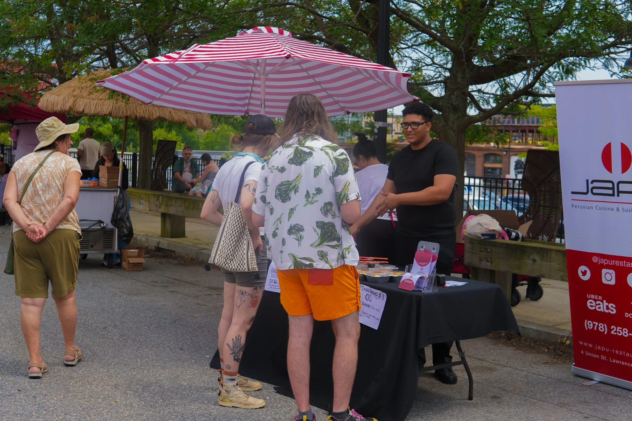 People at an outdoor food stall with a Japanese restaurant banner, under umbrellas, in a park-like setting during daytime.