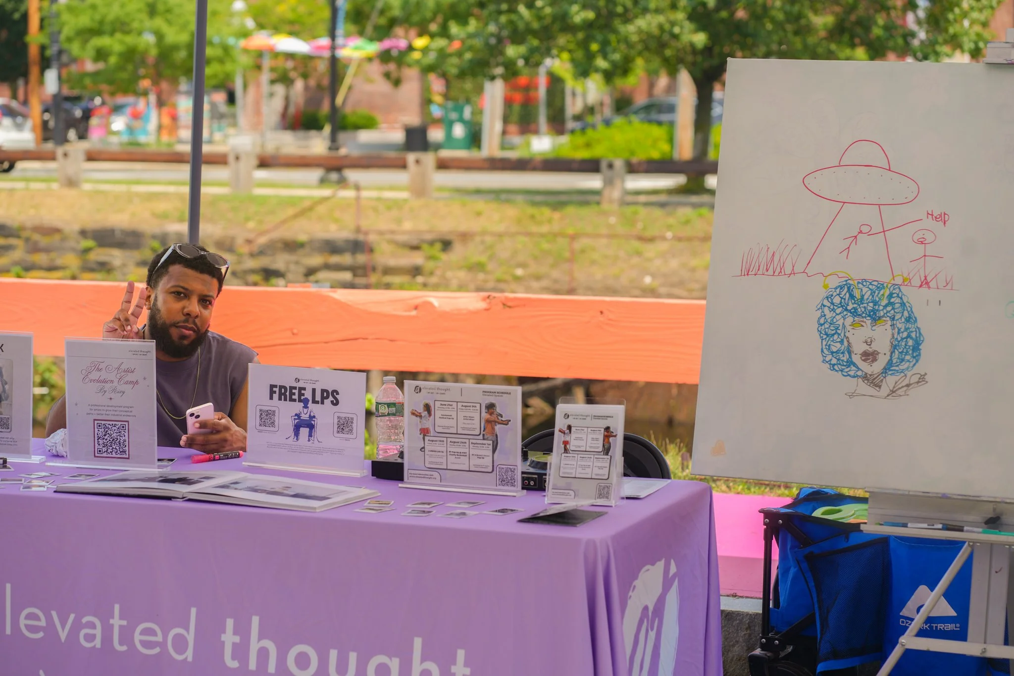A man sitting at a table with informational signs and pamphlets, making a peace sign with his fingers, at an outdoor event with trees and buildings in the background.