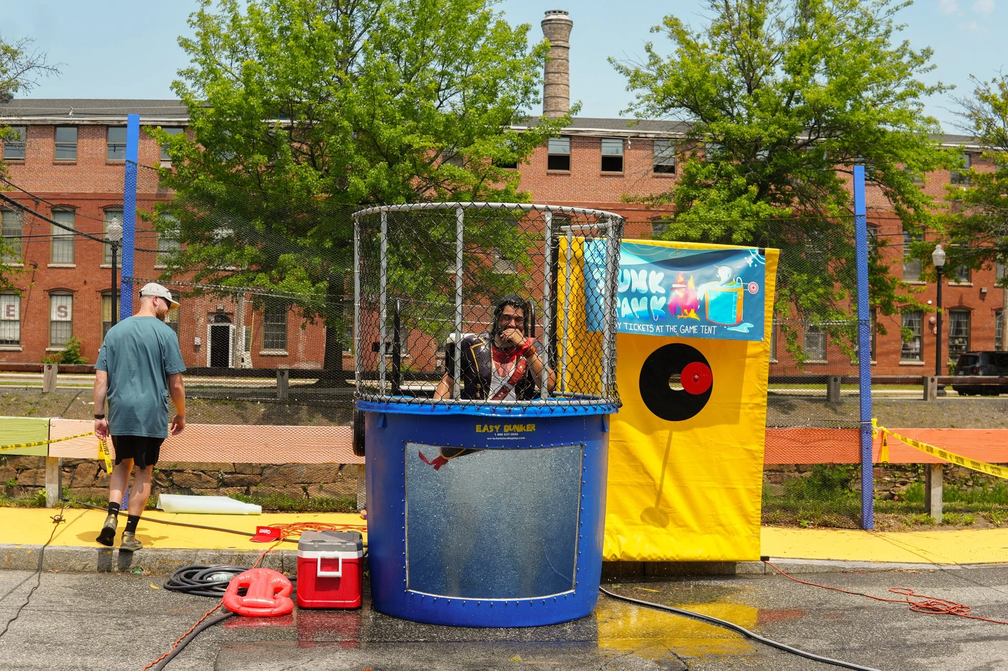 A person playing dunk tank at an outdoor event, with a man walking by, a yellow game tent, green trees, and brick buildings in the background.