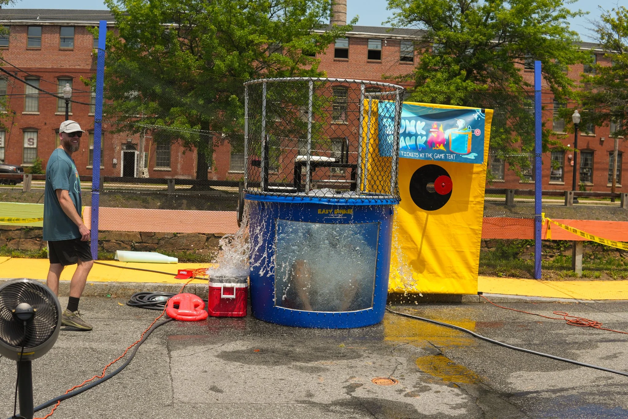 A man in a blue shirt, black shorts, and a white cap is standing next to a water dunk tank at an outdoor event, with water splashing inside. Behind him, there is a yellow and blue game tent with a black circle target and a brightly colored banner nex