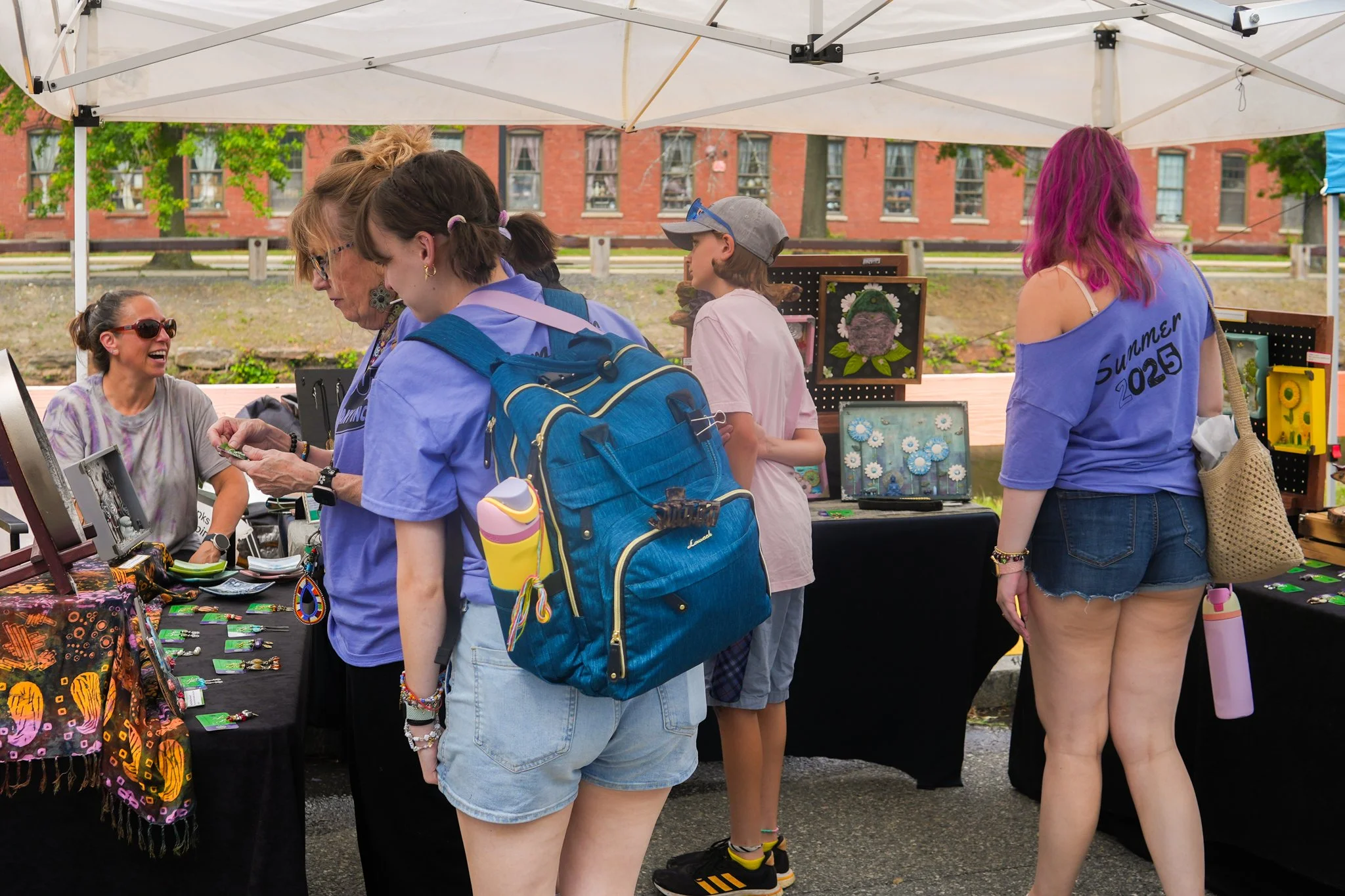 People shopping at an outdoor craft fair under a white canopy, with artwork displayed on tables.