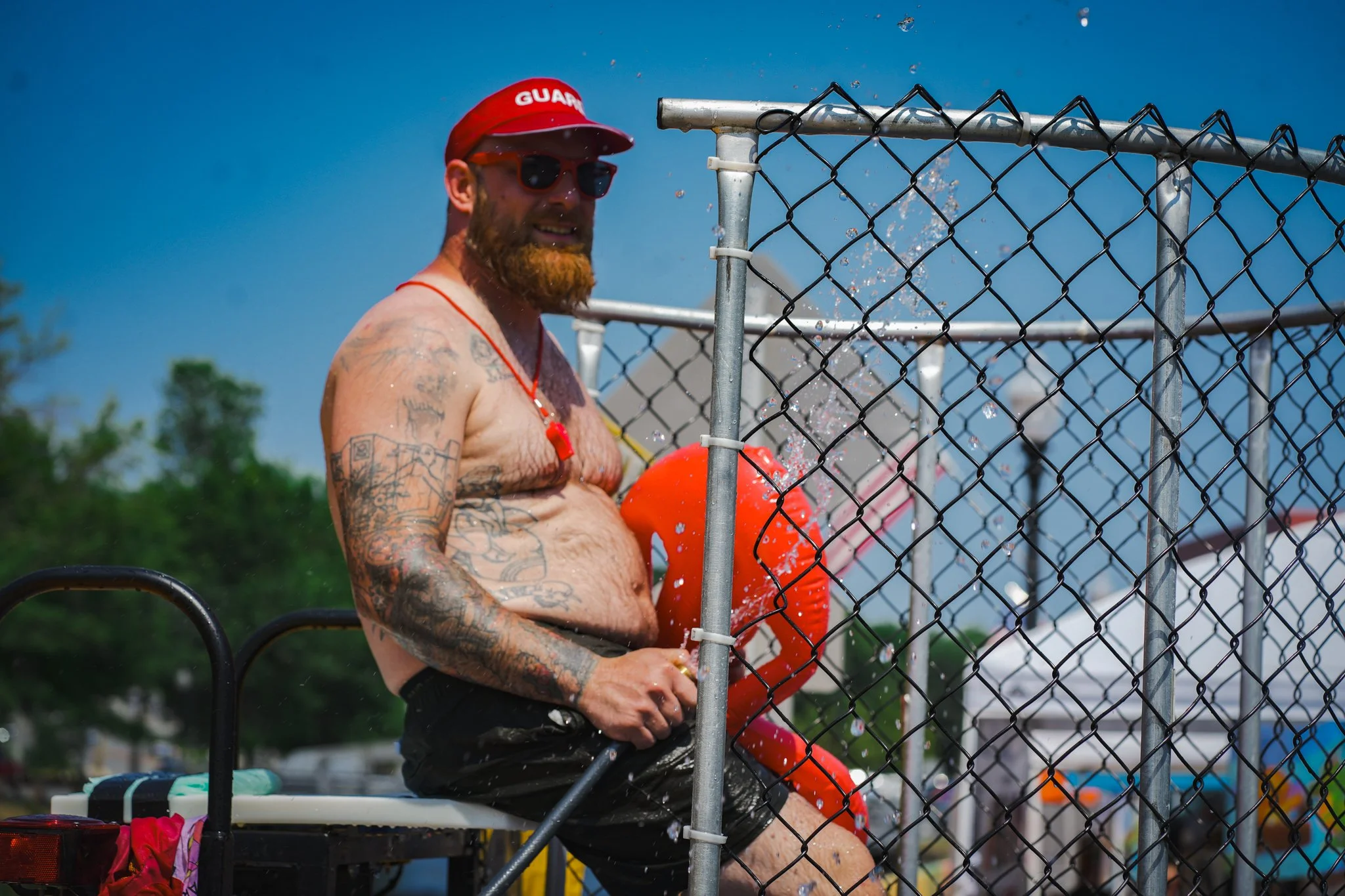 A shirtless man with tattoos, wearing sunglasses, a red cap, and a red whistle, sitting by a swimming pool with a water slide in the background, holding a red floatation device.