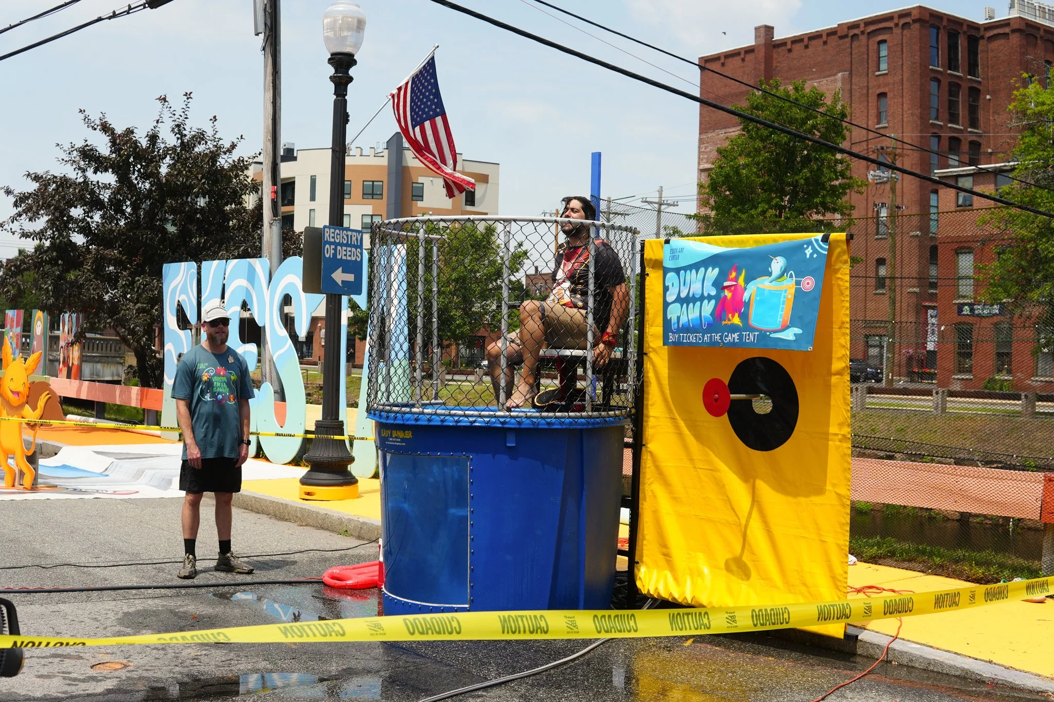 A man sitting on a dunk tank chair during a fair, with a banner reading 'Dunk Tank' nearby. A person stands outside the tank, and there are signs and caution tape around the area.