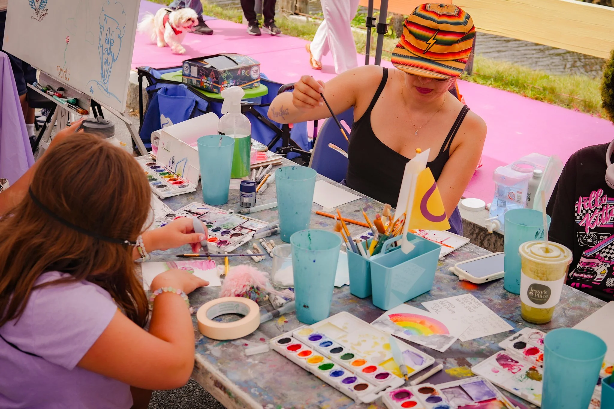 A woman and a girl are sitting at a table outdoors engaged in painting with watercolors, surrounded by art supplies including brushes, paint, and paper, near a pink mat and a whiteboard. The woman is wearing a colorful striped cap and black tank top,