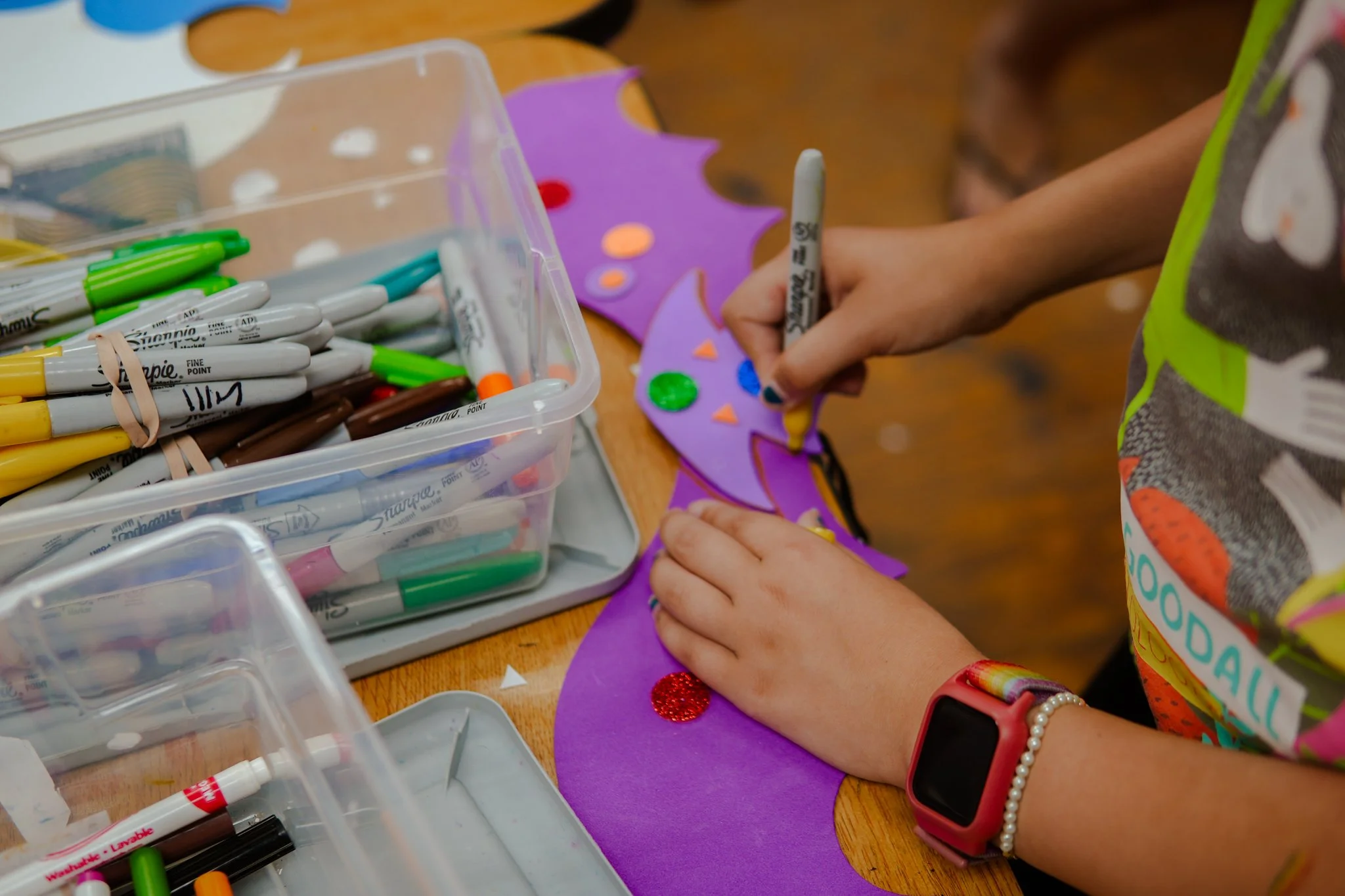 Child decorating purple paper with colorful stickers using a black marker, surrounded by containers of markers and craft supplies on a wooden table.