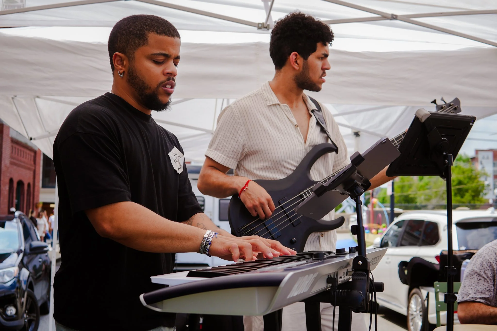 Two young men perform music outdoors under a white canopy; one is playing a keyboard, and the other is holding a bass guitar, both focused on their instruments.