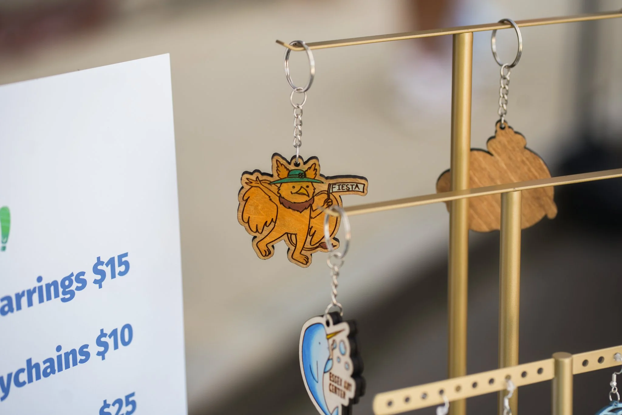 Wooden earrings shaped like a lion with a hat and a banner that says 'Fiesta,' hanging on a gold display stand.
