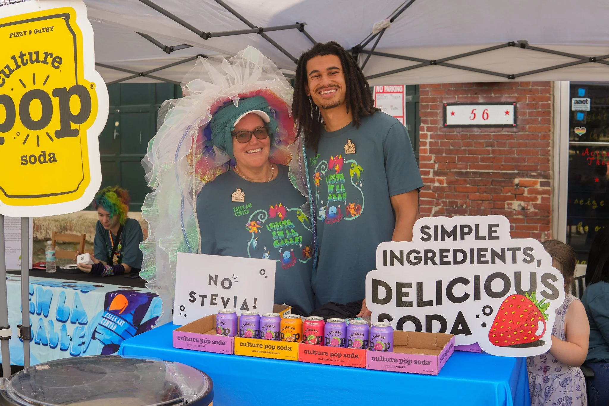 Two smiling people at a booth promoting Culture Pop Soda, with signs saying 'No Stevia!' and 'Simple ingredients, delicious soda' in front of a brick building. One person is wearing a colorful, frilly hat and glasses, and both are wearing dark blue T