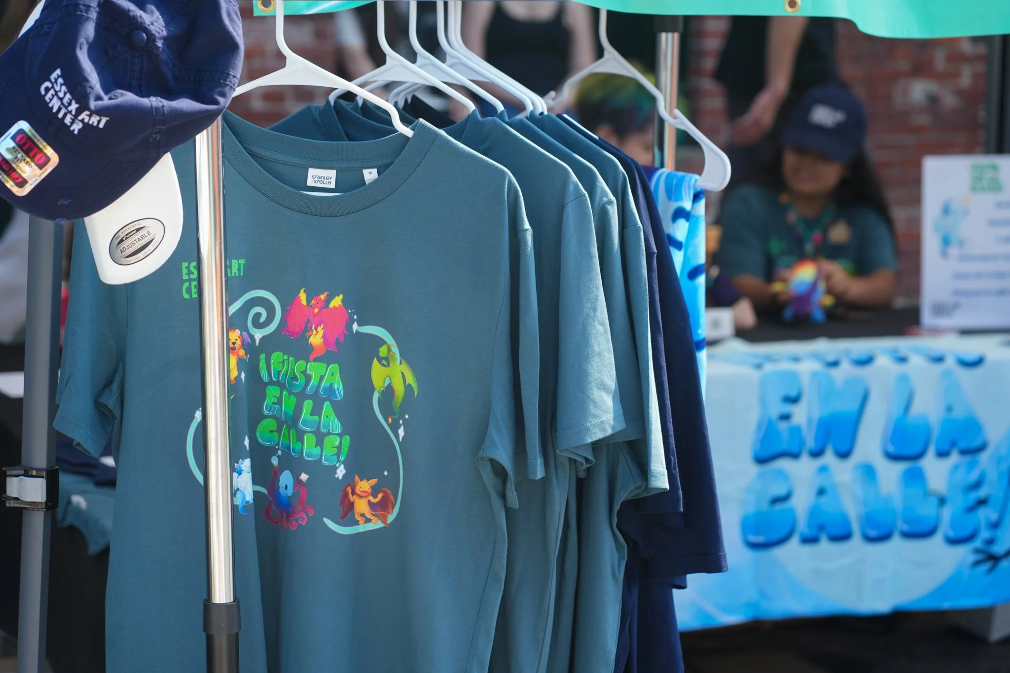 Display of blue t-shirts on a rack, with one featuring colorful graphic designs and text, at an outdoor event.