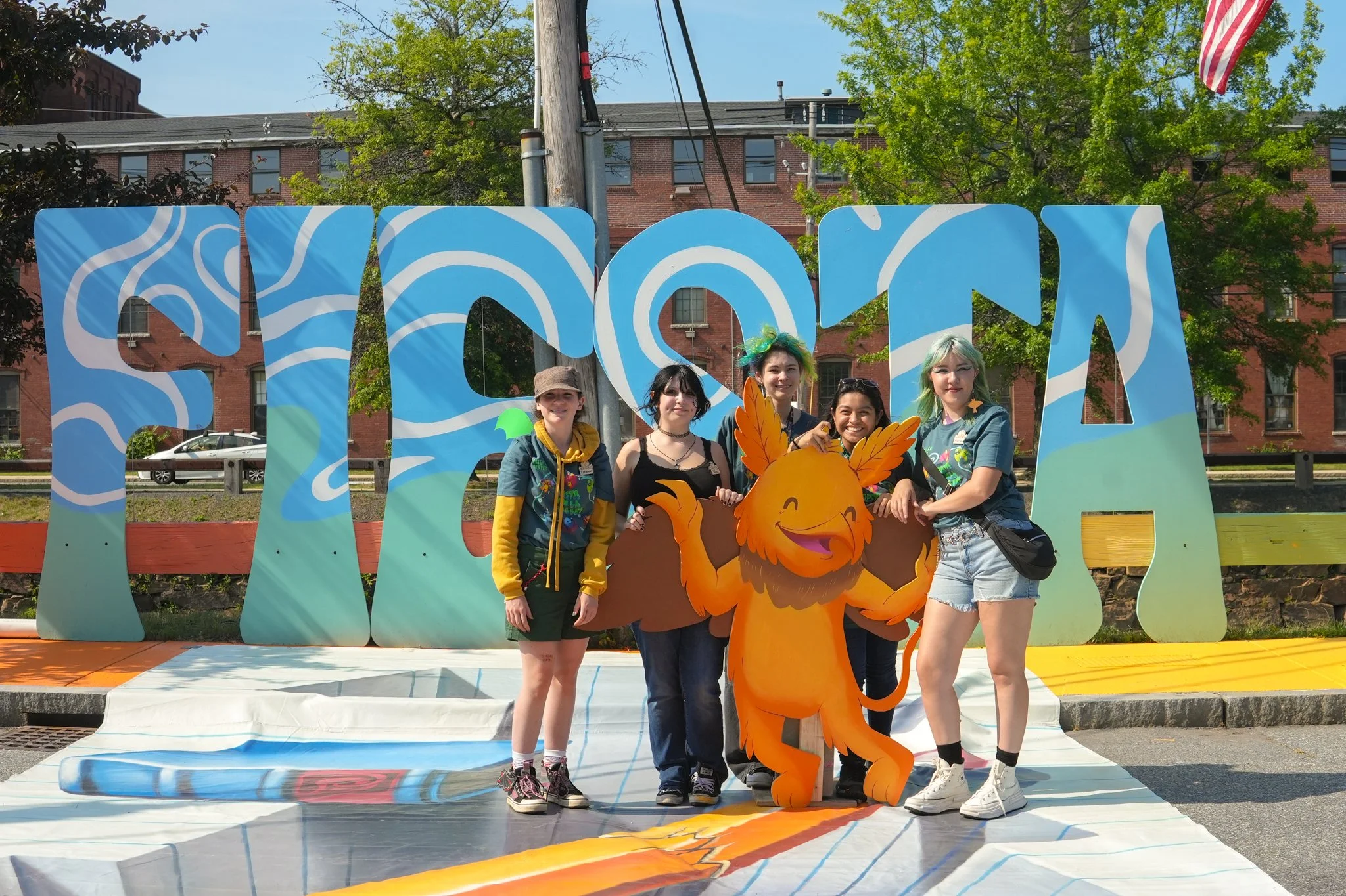Five young women standing in front of large colorful letters spelling 'SCOTA', with a cartoon lion mascot between them. They are outdoors on a sunny day, with trees and brick buildings in the background.