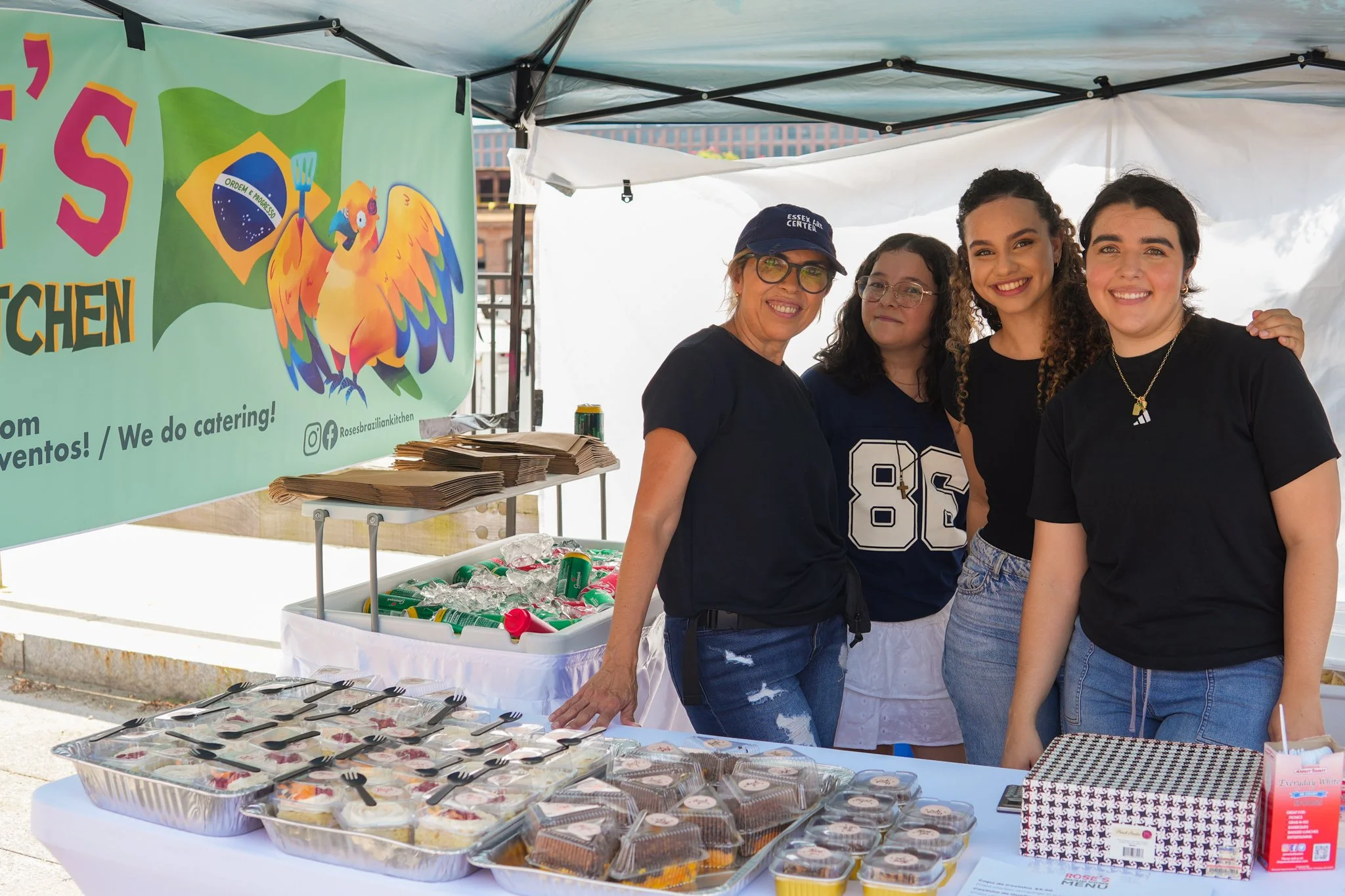 Four women standing together at a food stand with a large colorful banner behind them that features a parrot and the Brazilian flag. The table in front displays various cakes and desserts, with utensils and beverages nearby.