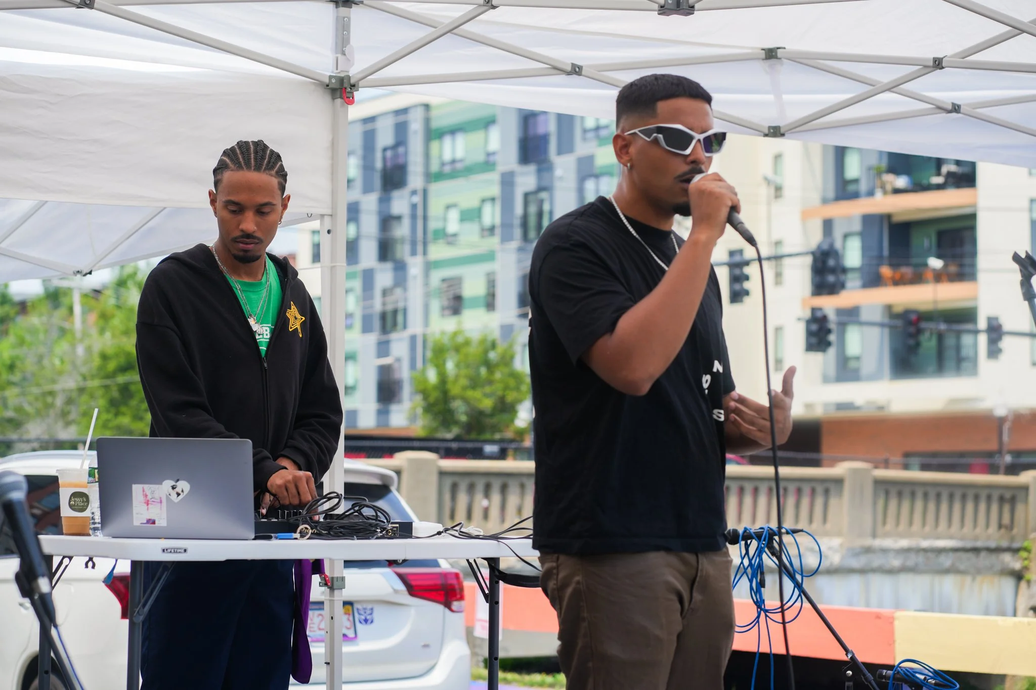 Two men, one with a microphone and the other with DJ equipment, performing outdoors under a white tent with city buildings in the background.