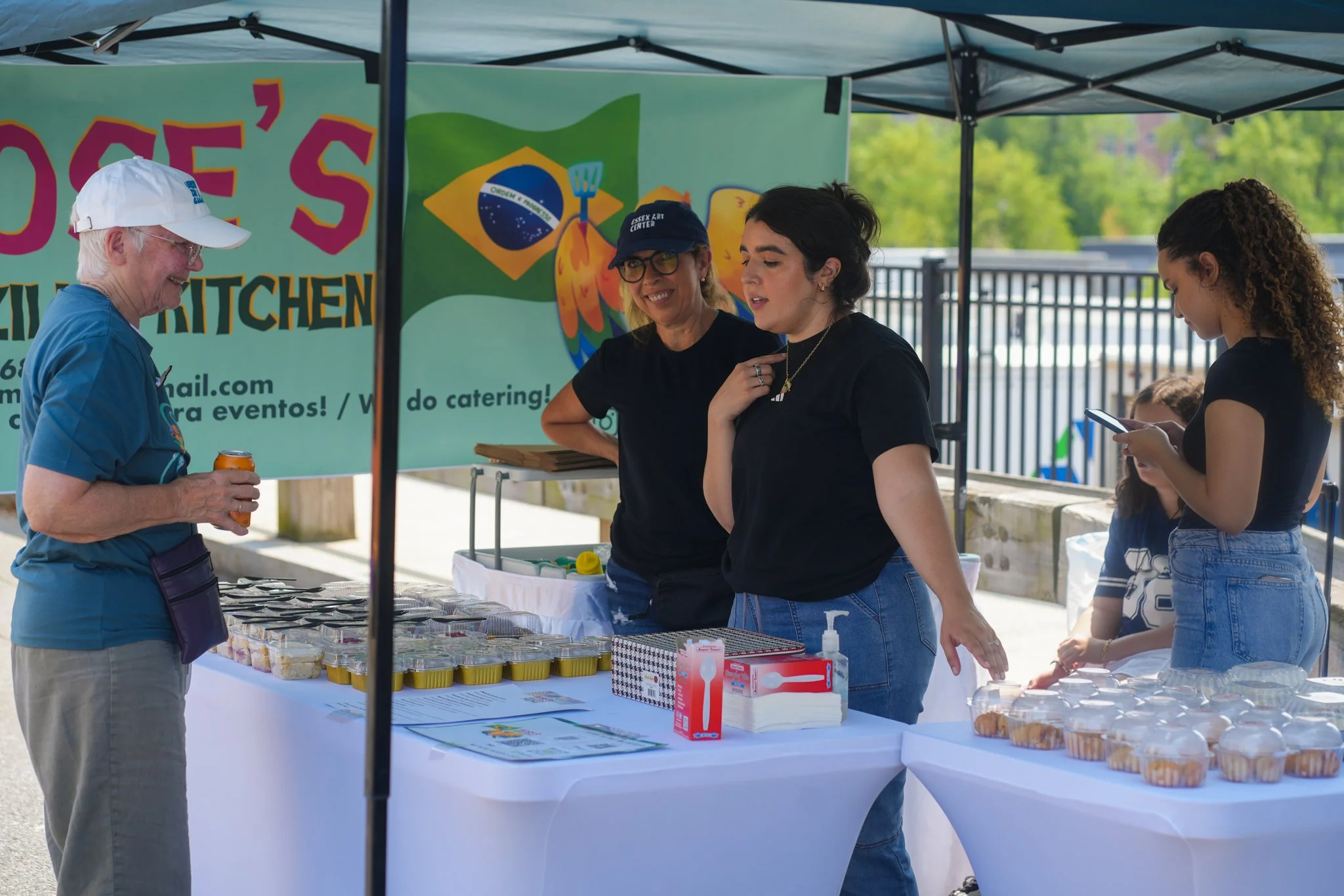 Four women at a food booth outdoors, with a banner reading 'Jose's Kitchen' in the background. One woman is holding a drink and smiling, another is wearing glasses and a cap, and two others are looking at their phones. The booth has containers of foo