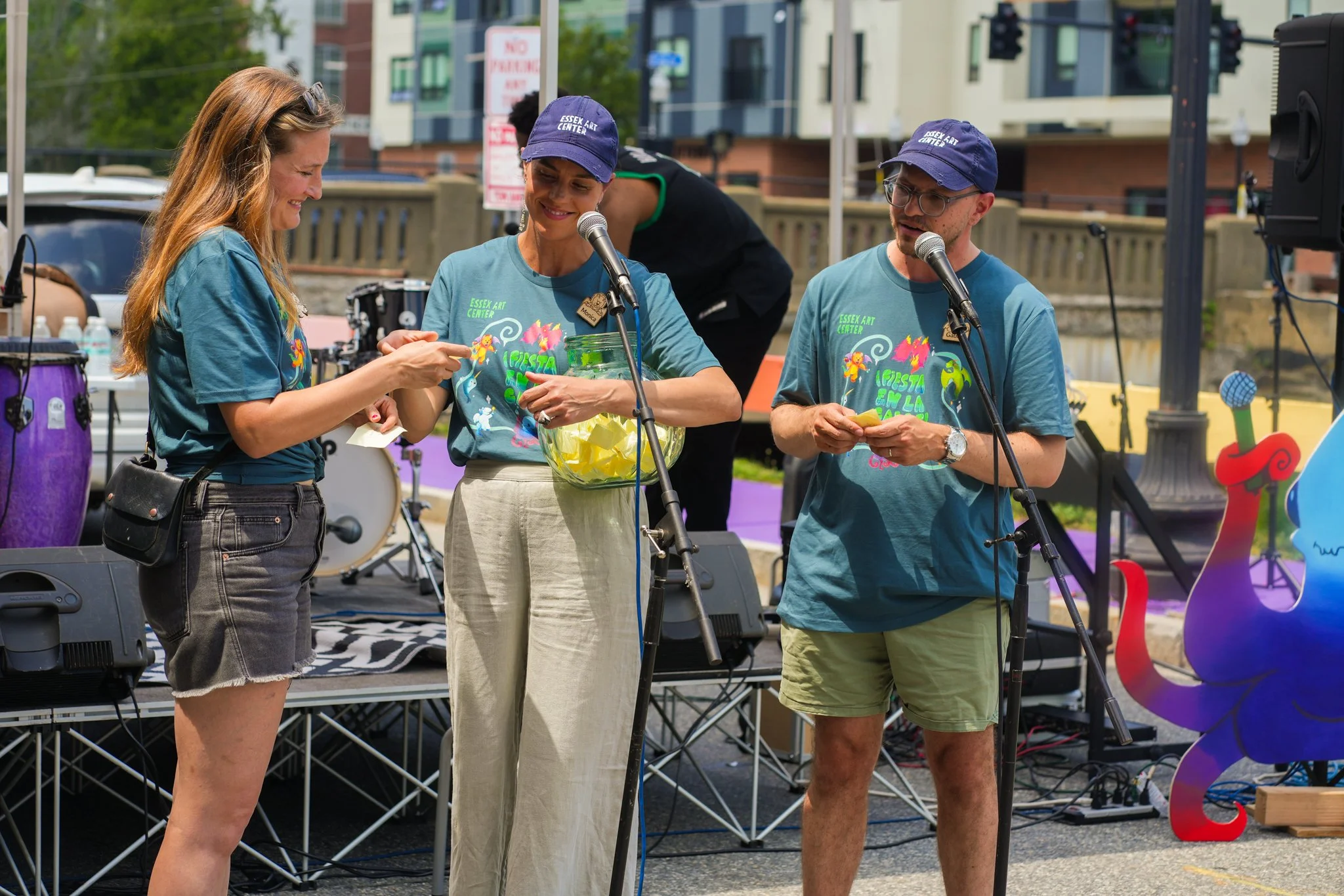 Three people standing on a stage during an outdoor event, two women and one man, with microphones, colorful decorations, and musical instruments in the background.