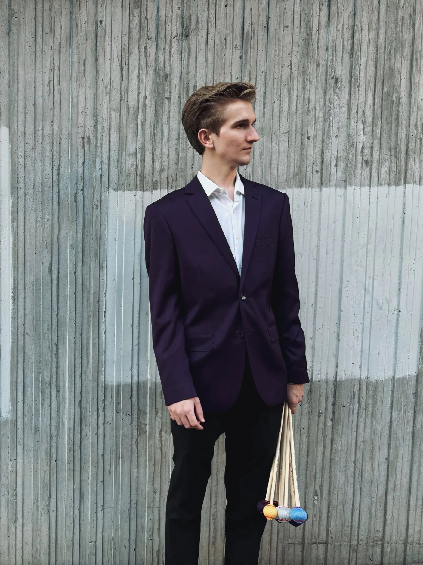 A young man in a navy blazer and white shirt standing against a concrete wall, holding mallets in his left hand.