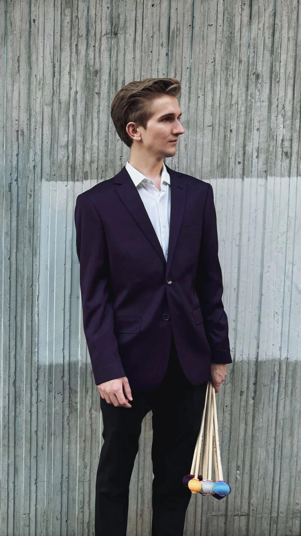 A young man in a dark suit and white shirt standing in front of a textured concrete wall, holding a bundle of colorful yarn balls.