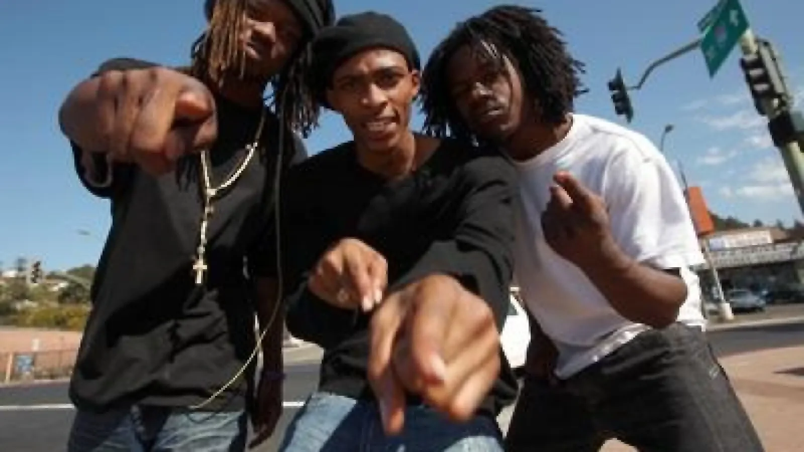 Three young men with dreadlocks posing on a city street under a clear blue sky. One in black shirt and black hat is pointing towards the camera, another in white shirt making a hand gesture, the third in black shirt with jewelry is leaning in.