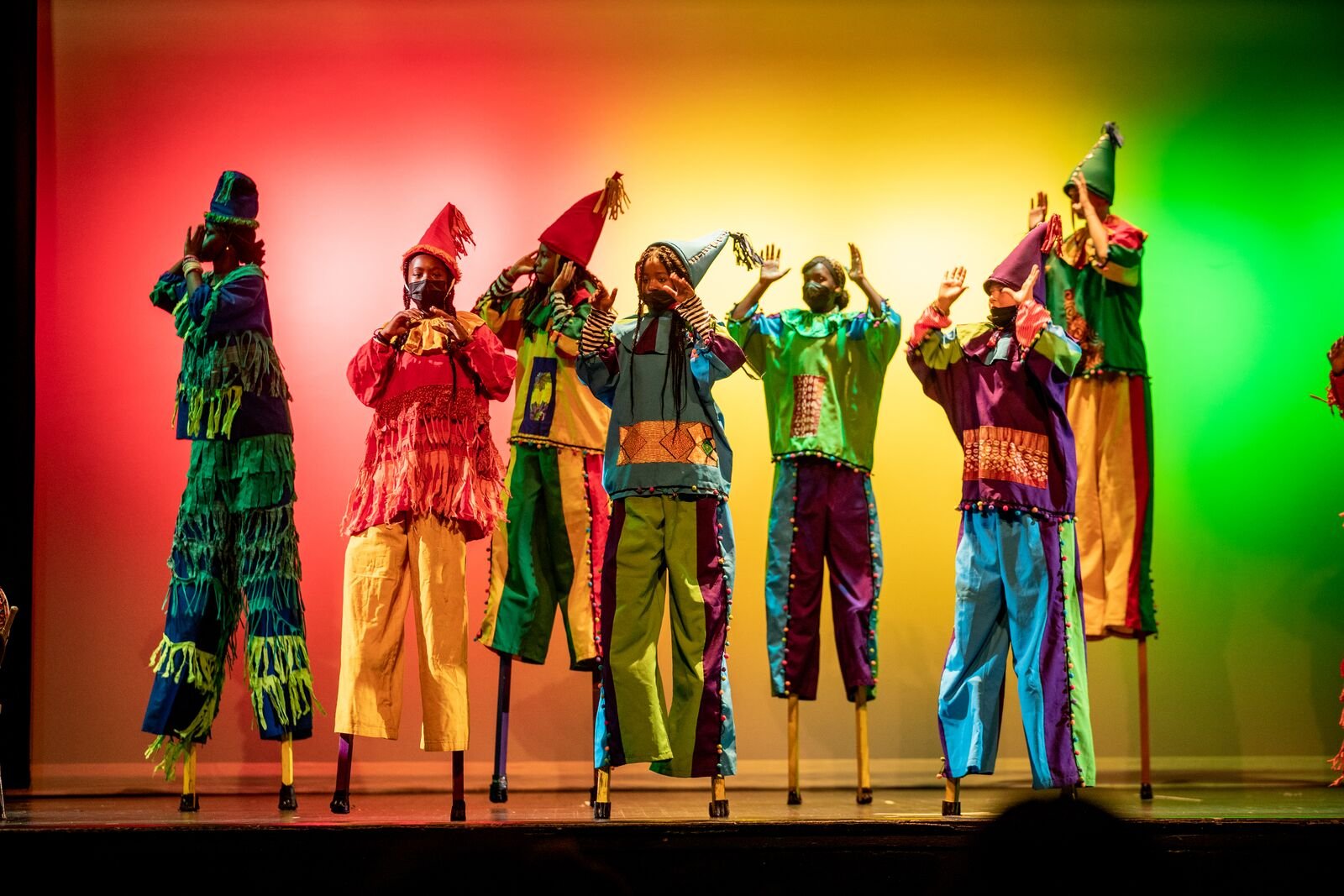Group of performers on stage dressed in colorful traditional African attire, wearing masks and hats, with a vibrant red, yellow, and green background.