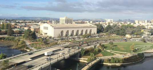 A city view with a large white building with arches, The Henry J. Kaiser Center for the Arts, a highway with cars, and a park with green grass and trees under a partly cloudy sky.