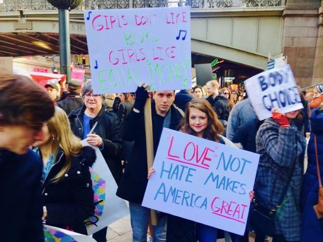 Sarah Dubow, health coach and wellness expert, and partner Nick holding up signs at women's march that say "Girls don't like boys, girls like equal money" and "love not hate makes America great"