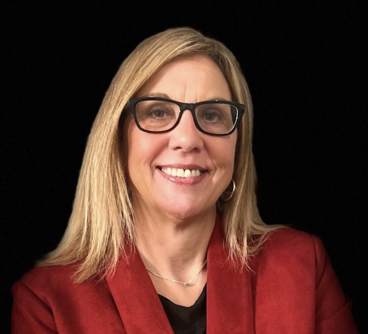 A woman with shoulder-length blonde hair, wearing black glasses, a red blazer, and a black top, smiling against a black background.
