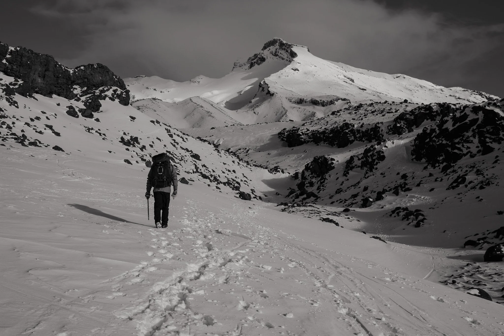 A mountaineer hiking in snowy mountain terrain with a backpack and walking stick, snowy peaks in the background under a cloudy sky.