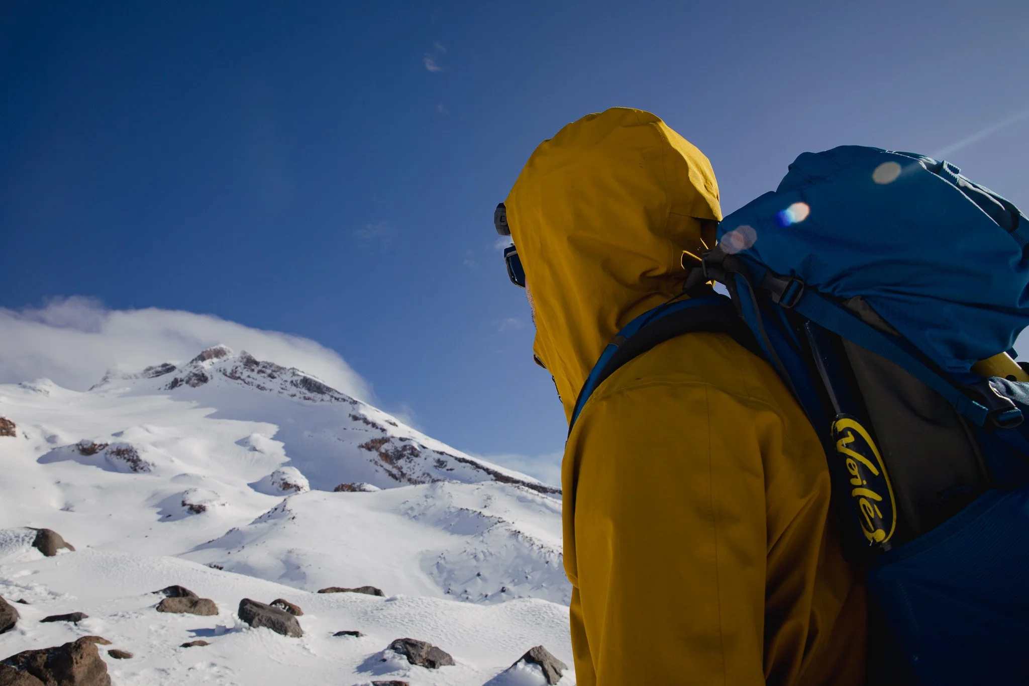 A person wearing a yellow jacket and a blue backpack standing in a snowy mountain landscape, with a snow-covered peak and a blue sky in the background.