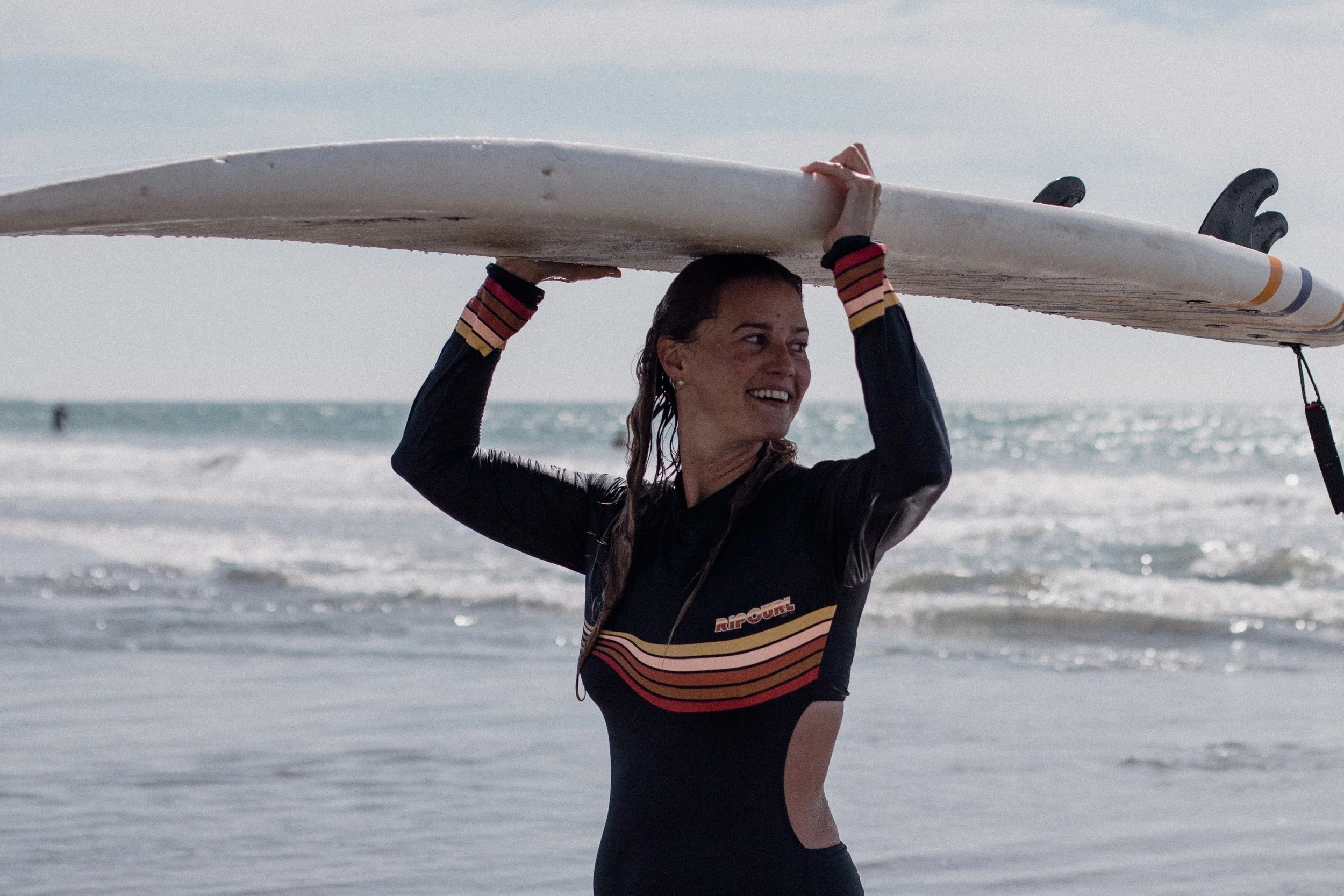 A woman in a black wetsuit holding a surfboard above her head on a beach with ocean waves in the background.