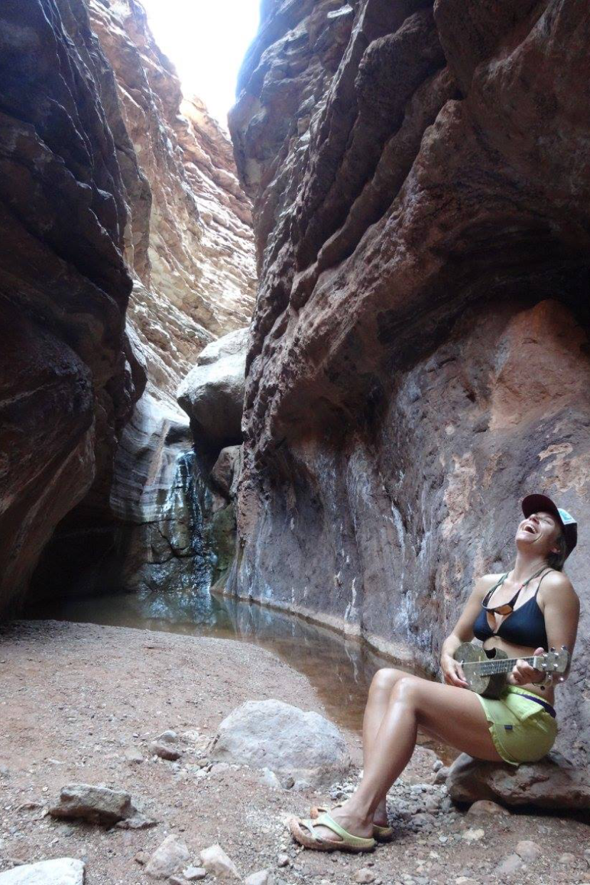 A woman sitting on a rock at the bottom of the Grand Canyon, playing a hemp ukulele and smiling joyfully.