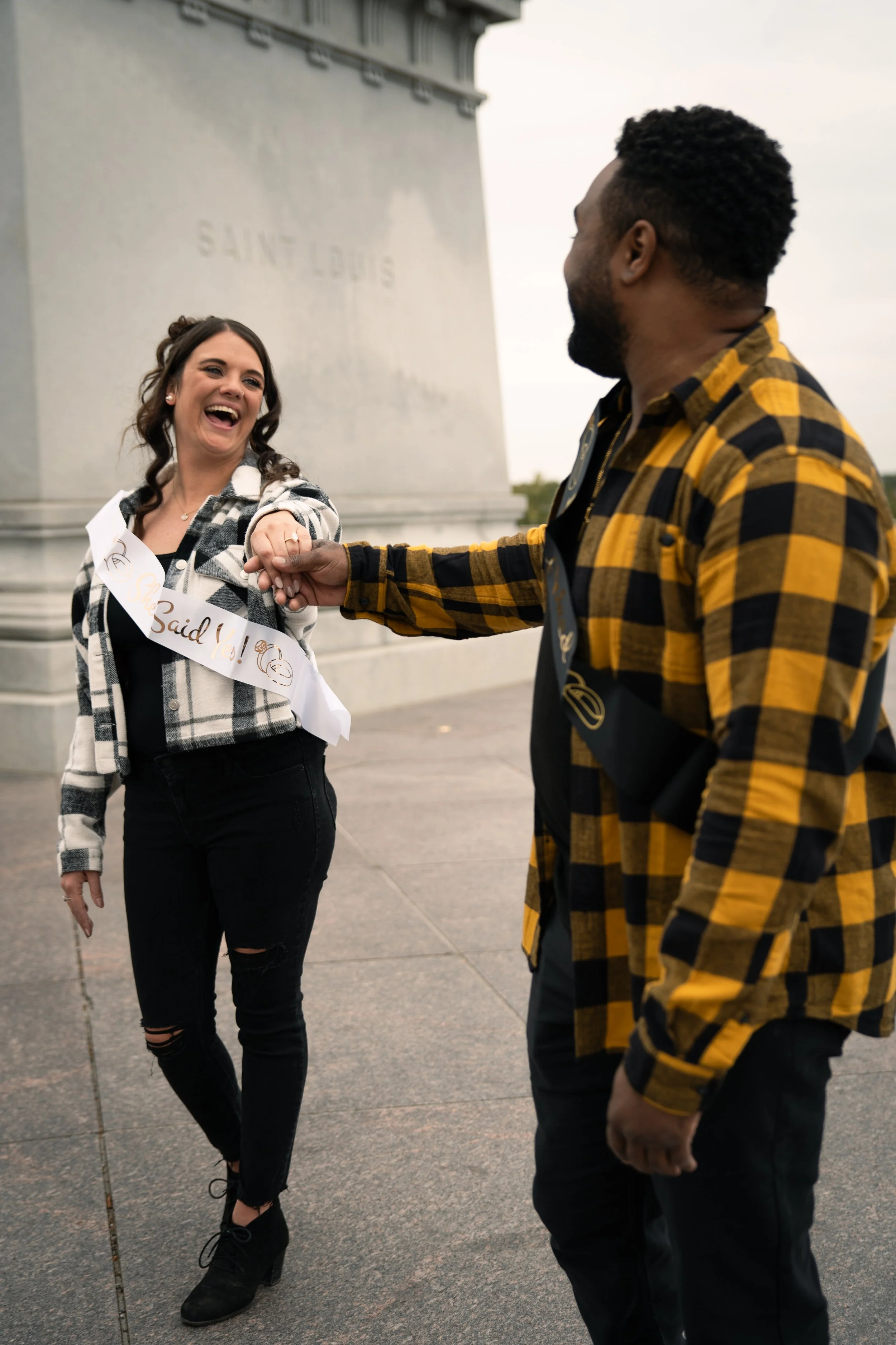 A woman and a man are smiling and holding hands in front of a monument, with the woman wearing a 'Said Yes!' sash.