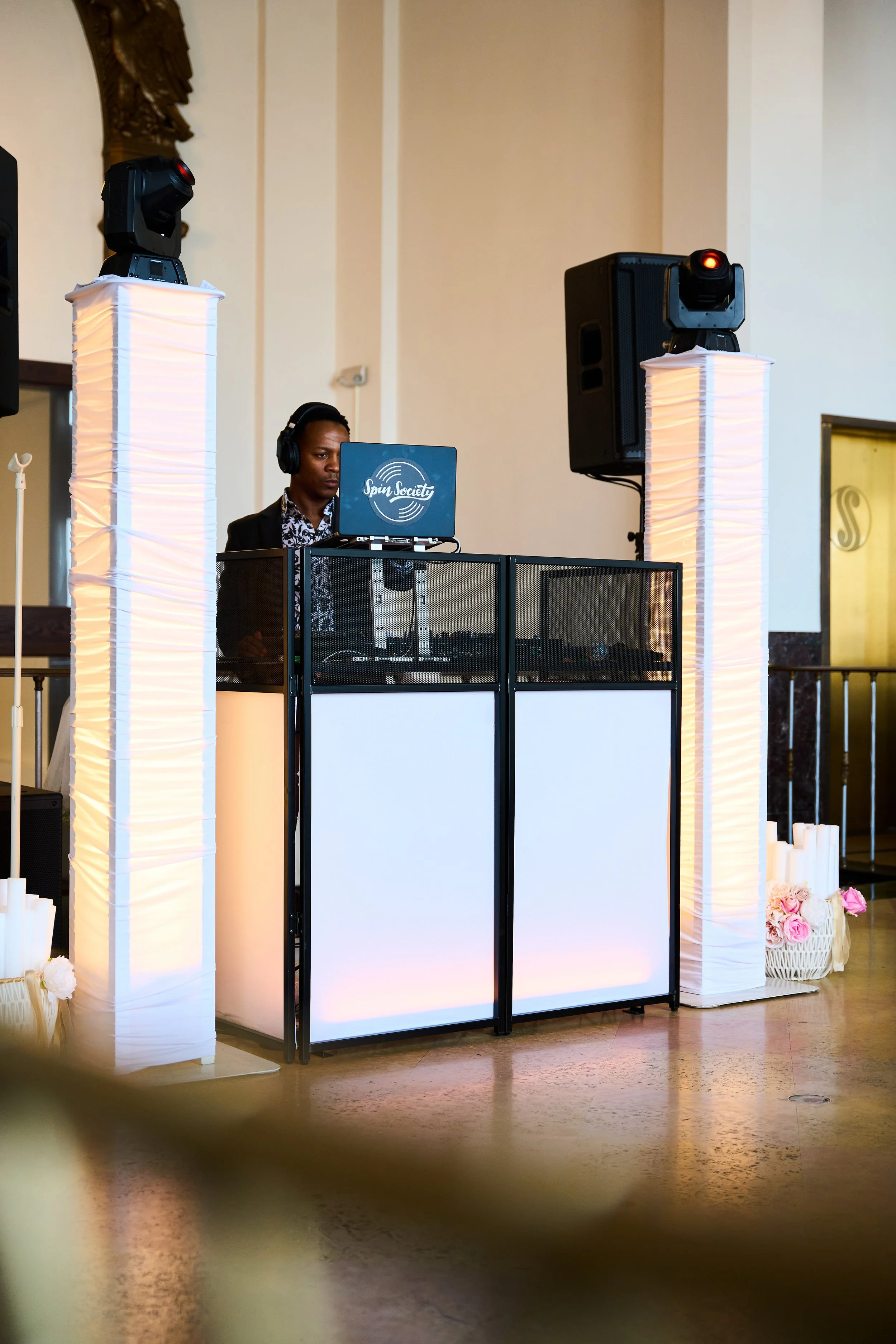 A DJ with headphones standing behind a DJ booth at an event, with decorative lamp columns and floral arrangements.