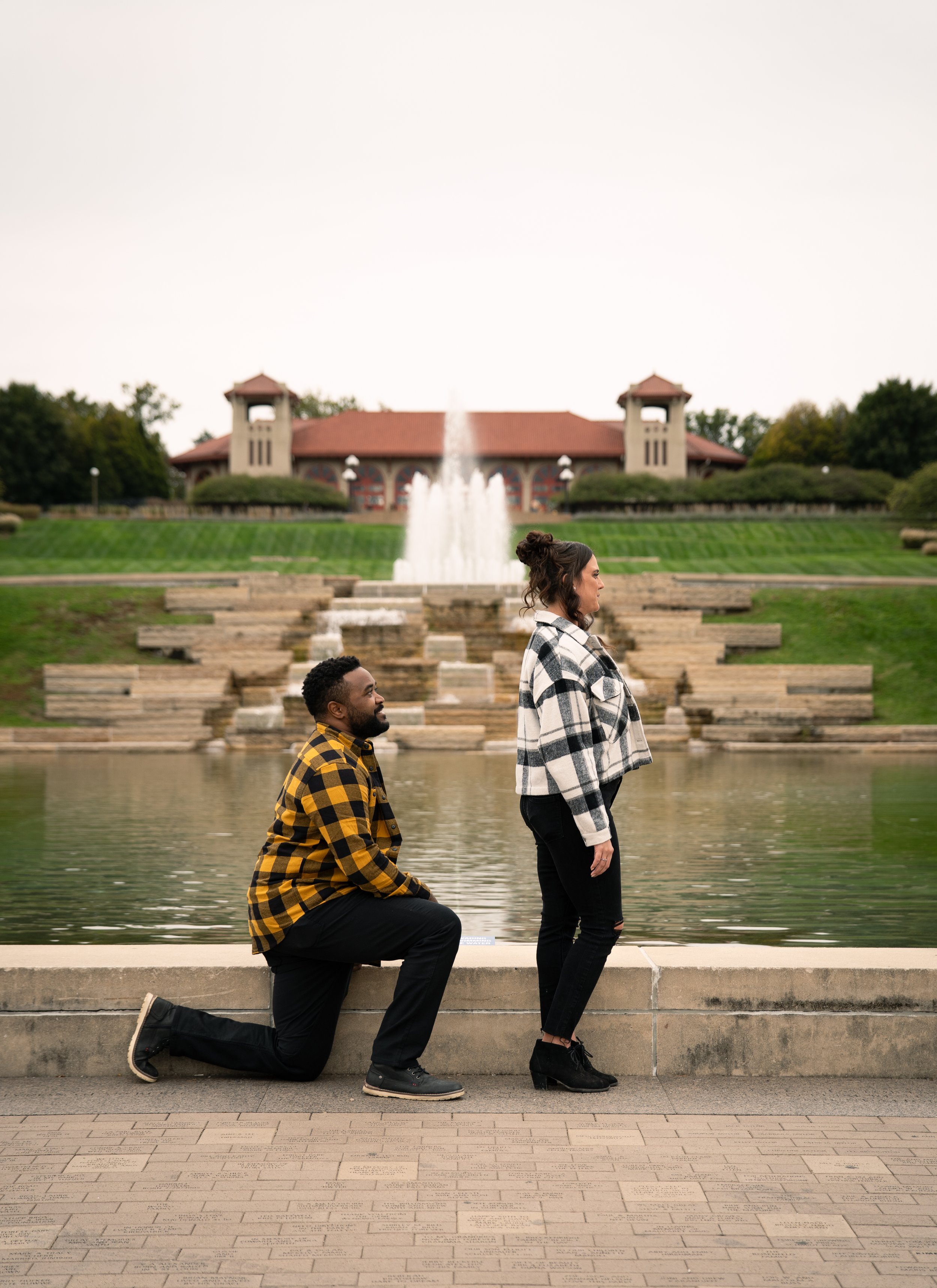 A man kneeling on one knee, proposing to a woman by a fountain in a park with a large building and fountain in the background.