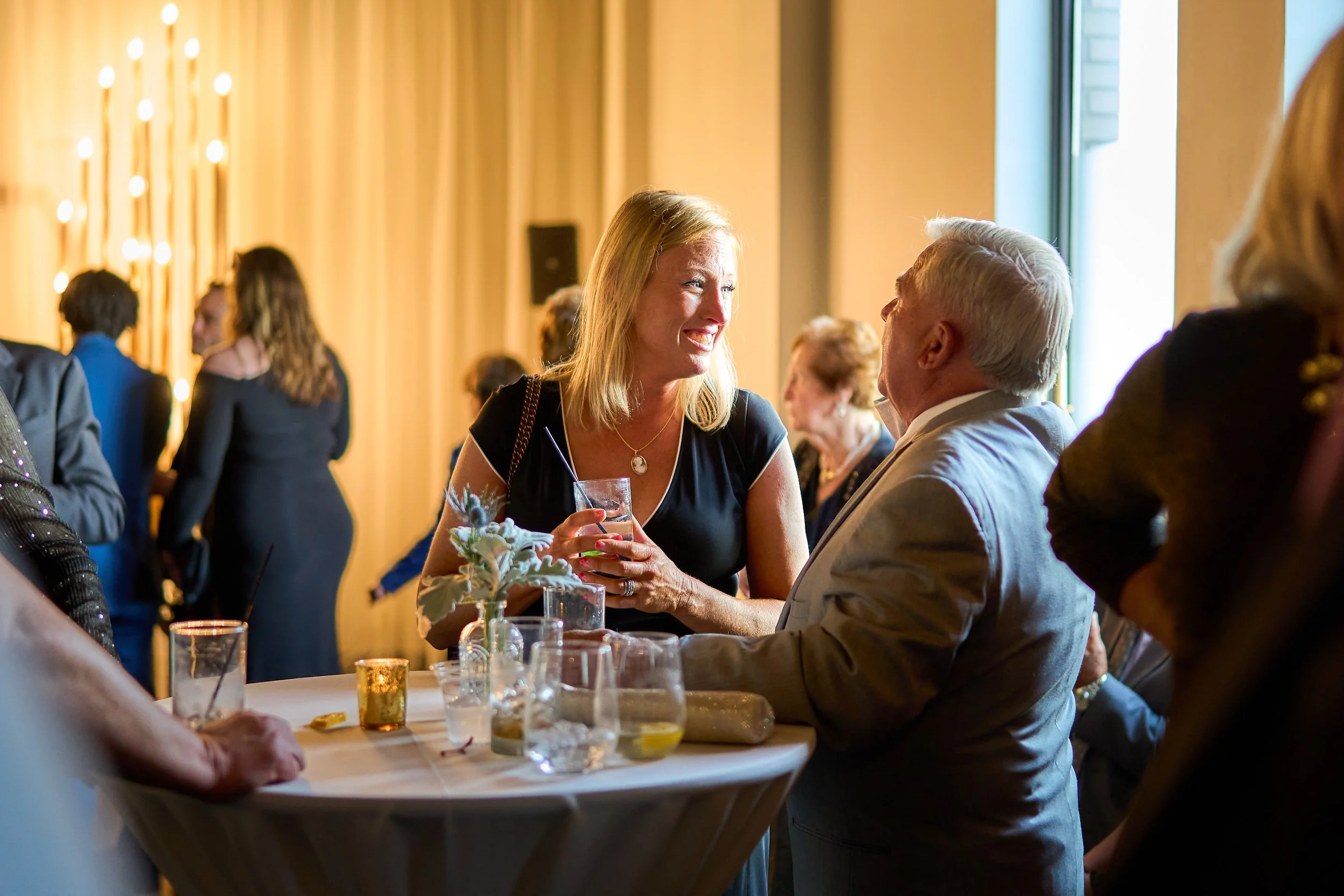 A woman with blonde hair in a black dress smiling and talking to an older man with white hair in a gray suit, at a social event with other people in the background.