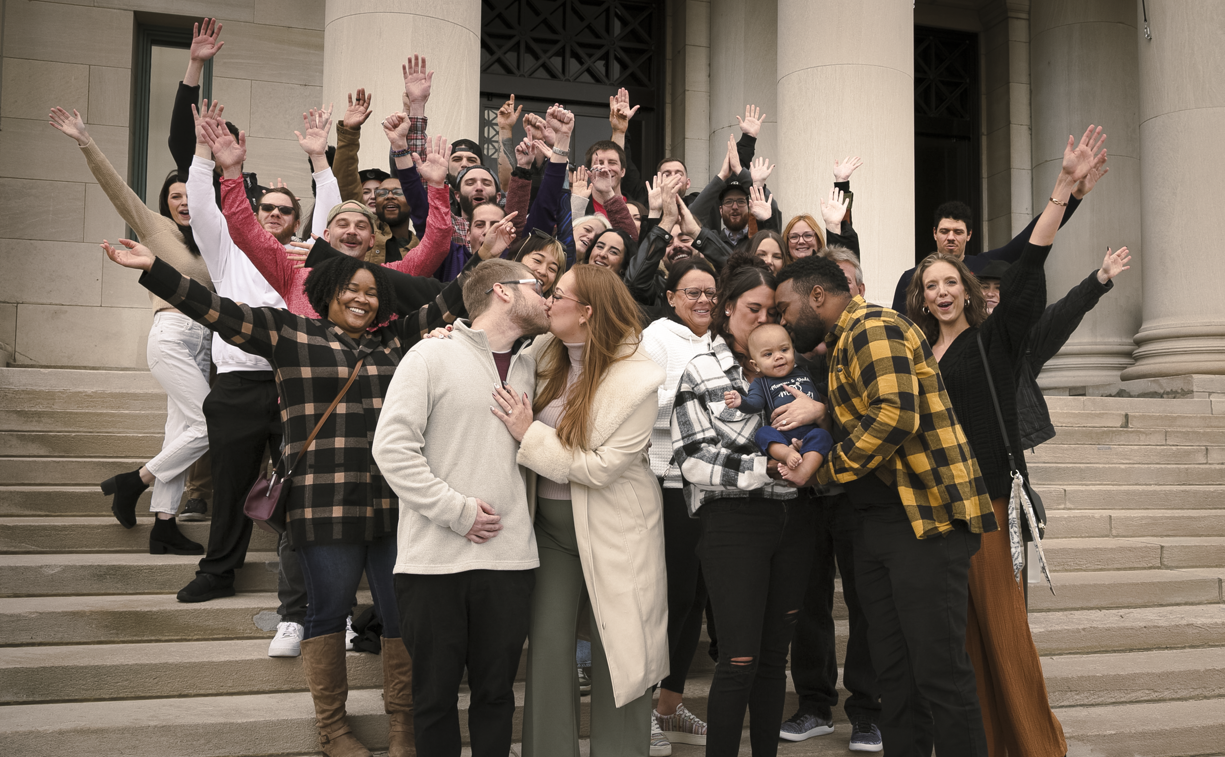 Group of diverse people on steps celebrating, with some kissing and holding a baby, in front of a building with large columns.
