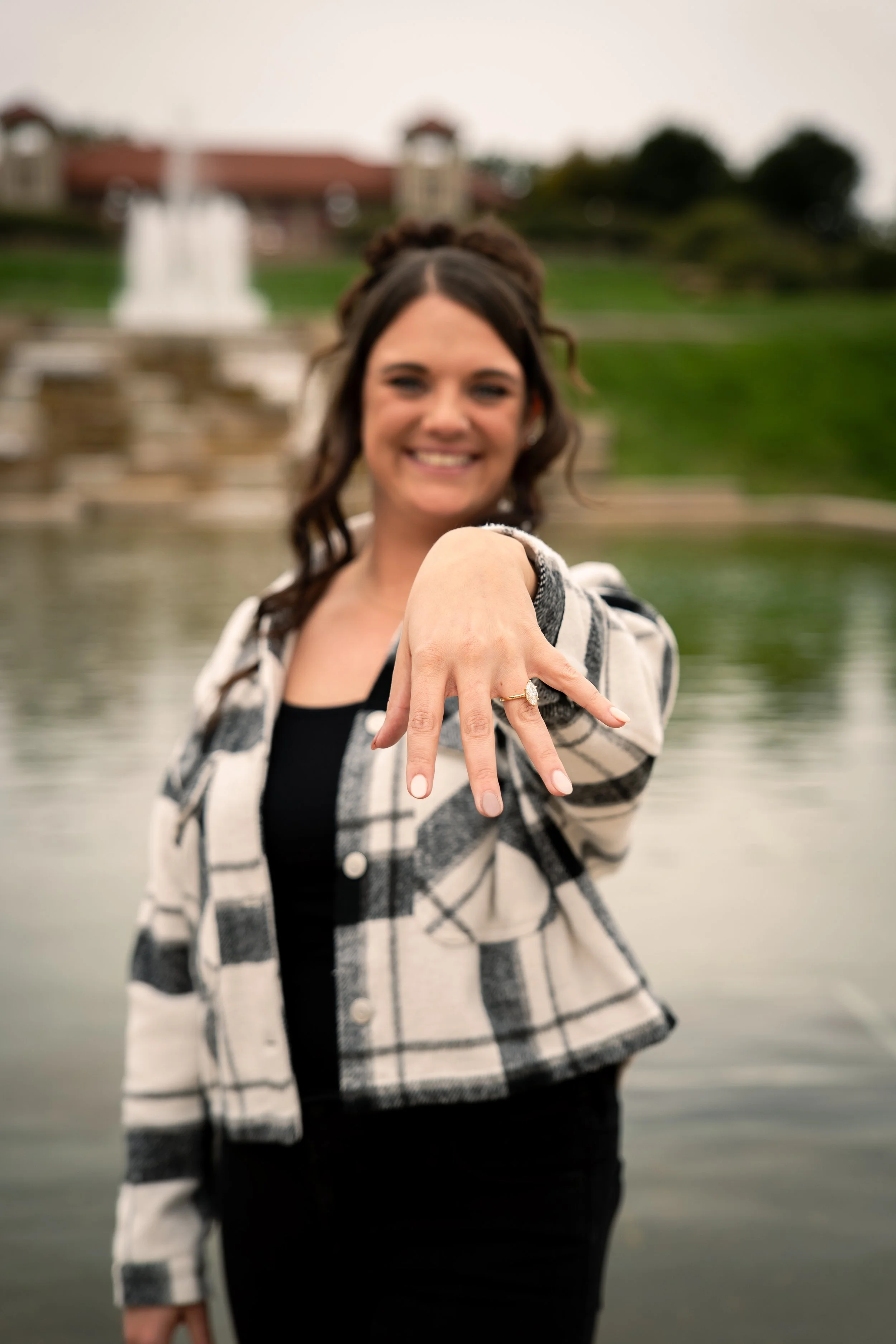 A woman smiling and showing her engagement ring on her left hand, with a blurred outdoor fountain and greenery in the background.