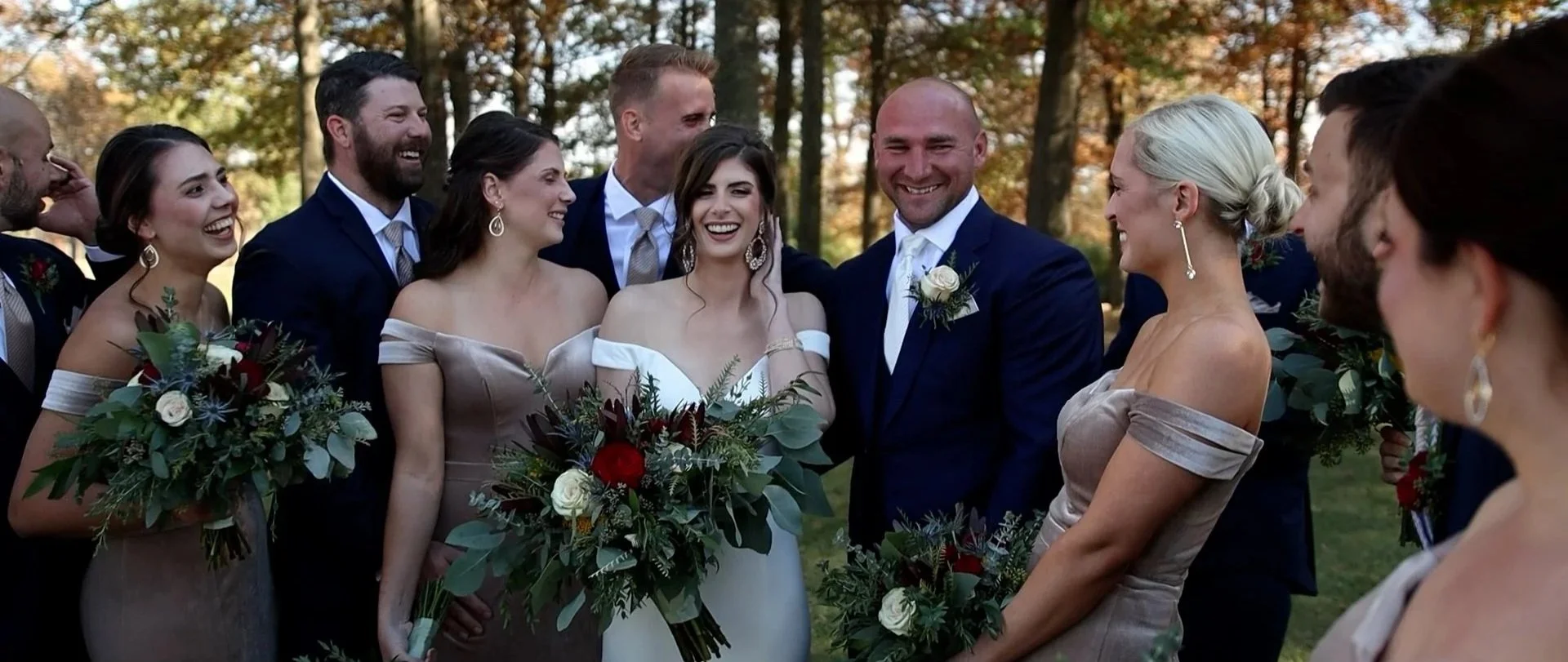Black and white photo of a wedding party with ten people, including the bride and groom, posing outdoors against a brick wall and string lights, with others holding bouquets of flowers.
