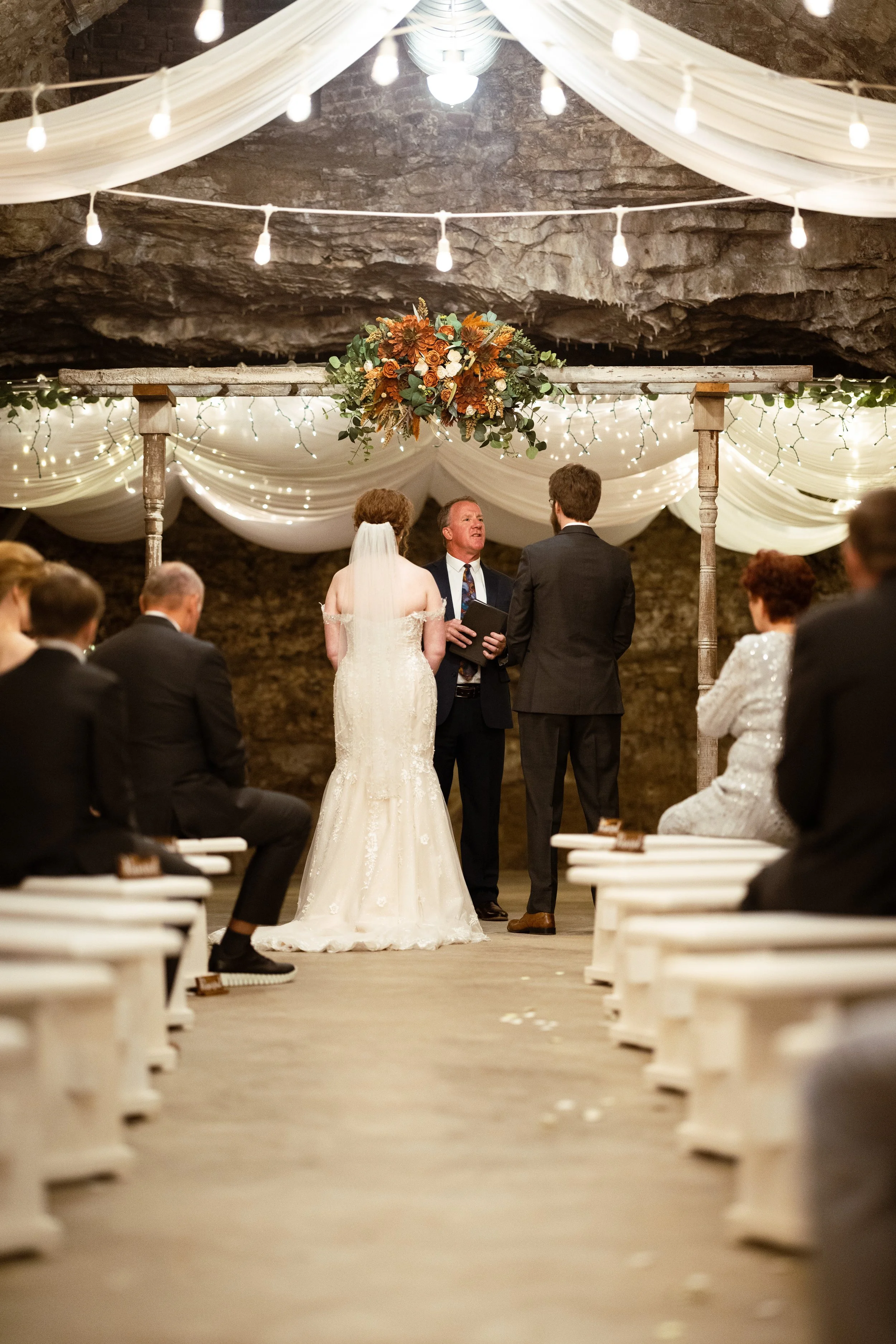 A wedding ceremony taking place in a cave with draped white fabric, string lights, and a floral arrangement overhead. The bride and groom stand at the altar facing an officiant, with guests seated on either side.