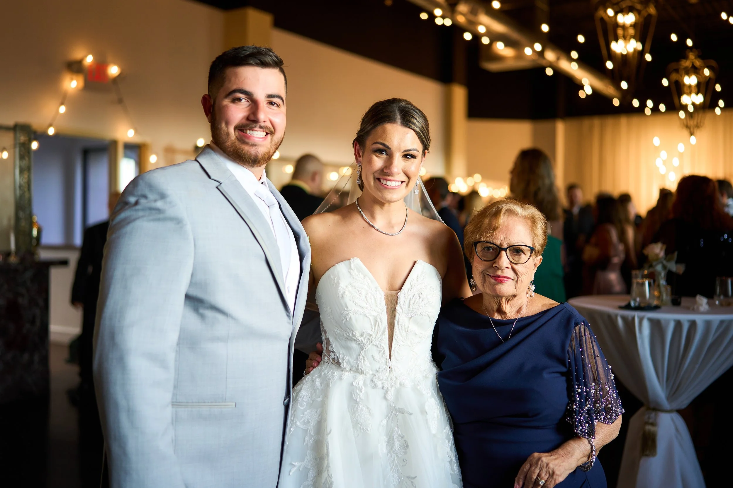 Three people smiling at a wedding reception, including a man in a light gray suit, a woman in a white wedding dress, and an older woman in a dark blue dress with glasses, in a decorated venue with string lights.