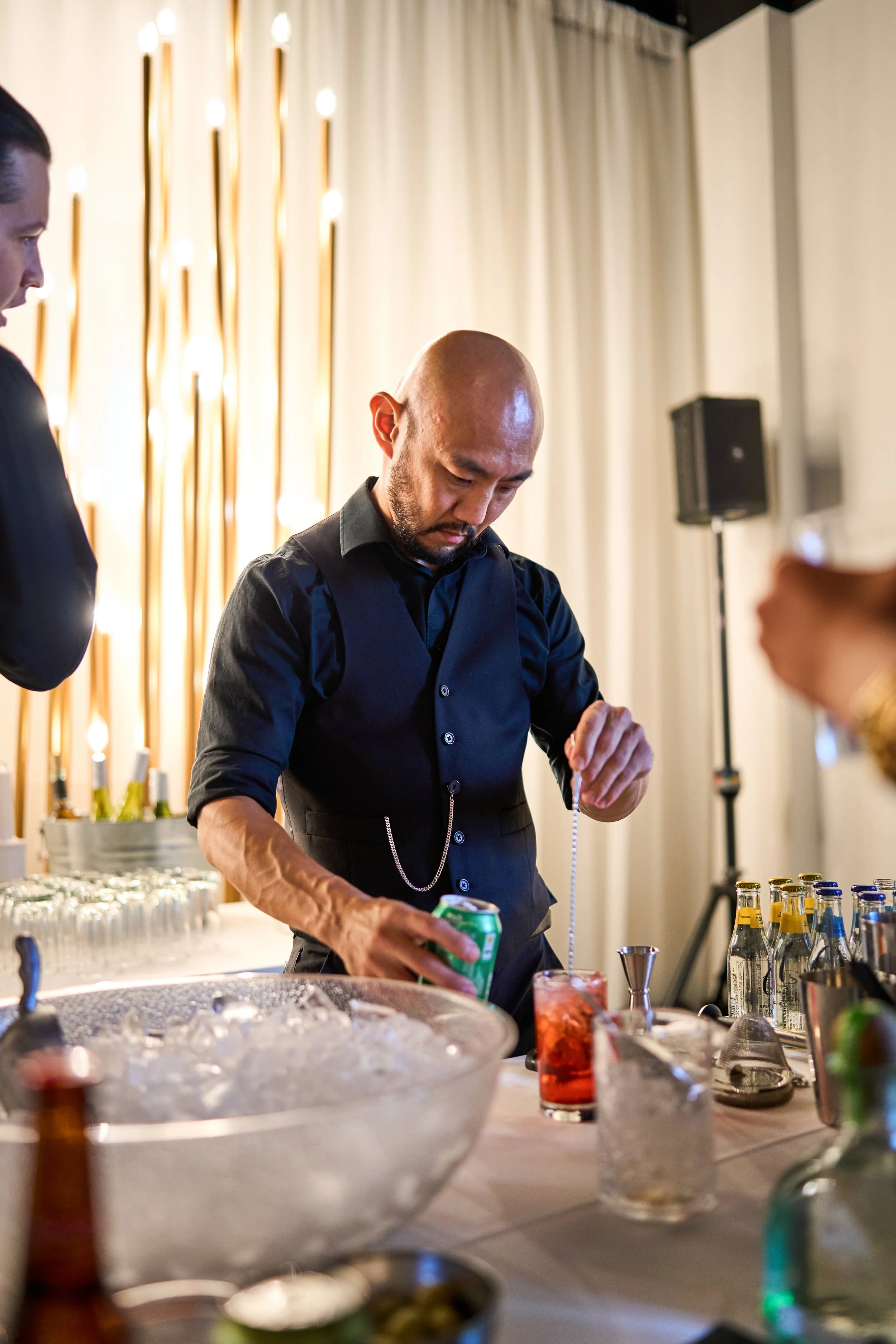 A bartender in black shirt preparing a cocktail at a bar, with various bottles and bar tools on the counter.