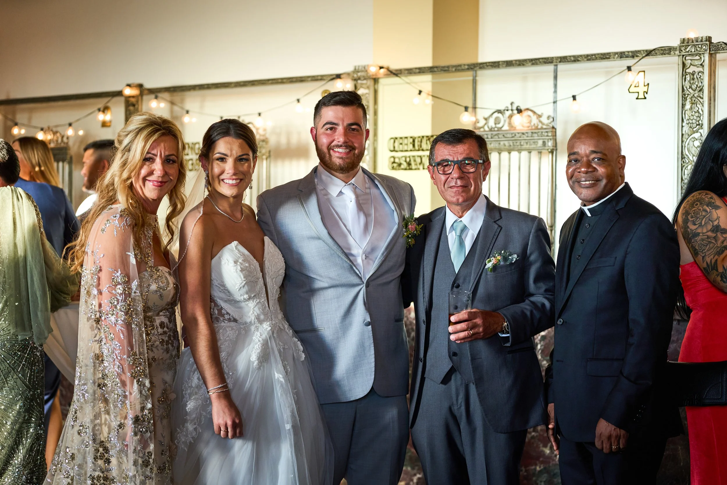 A wedding reception with six people dressed in formal attire, smiling for a photo. The bride wears a white gown, and the groom wears a gray suit. The others include two men and two women dressed elegantly, standing in a decorated indoor venue.
