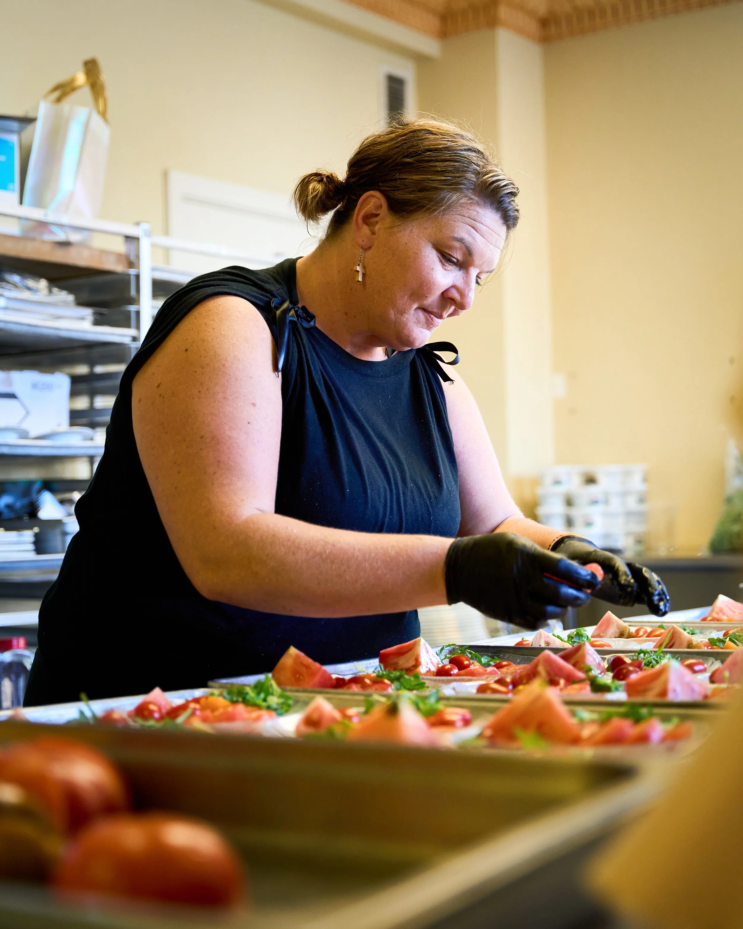 A woman wearing a black sleeveless top and black gloves is preparing food, garnishing plates with tomatoes and herbs in a kitchen or food prep area.