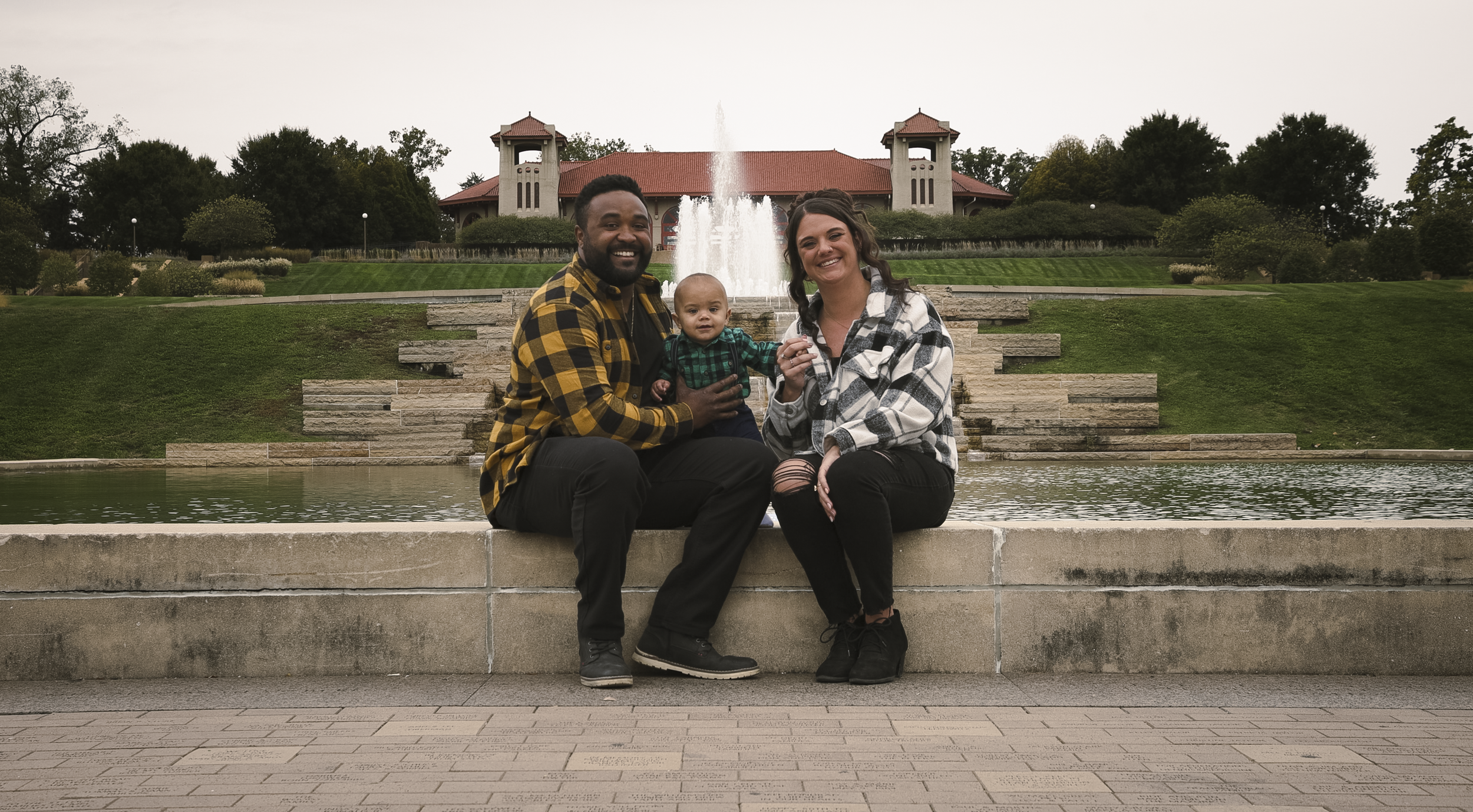 Smiling family of three sitting on a fountain ledge in a park with a building and fountain in the background. The man is wearing a yellow plaid shirt, the woman a black and white plaid shirt, and the child a green plaid shirt.