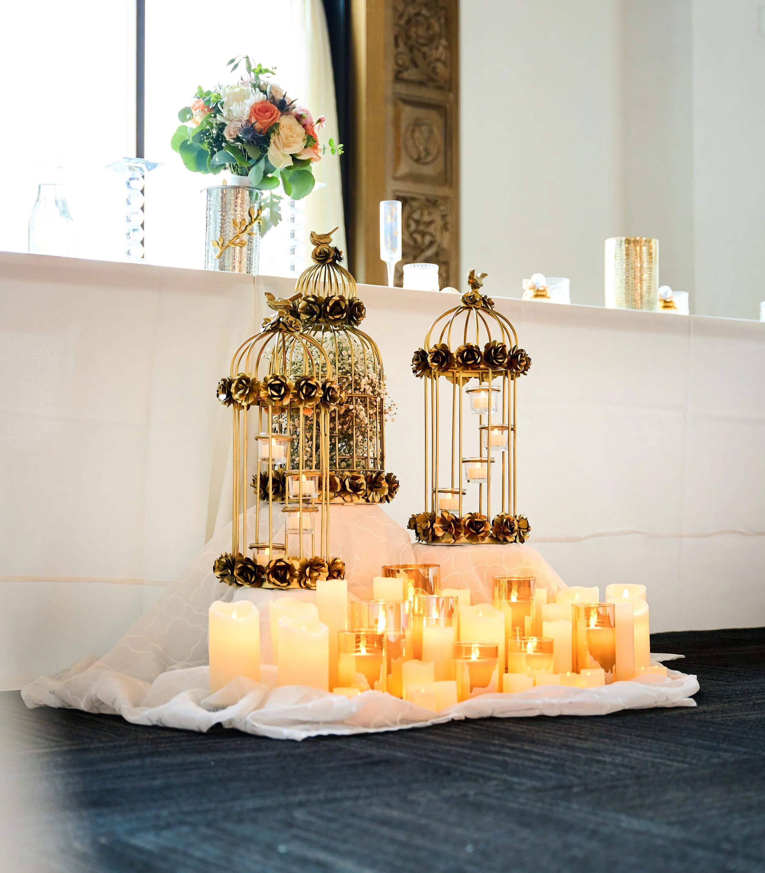 Decorative display featuring gold birdcages with flowers, surrounded by lit candles on a white fabric, with a vase of flowers and glassware in the background.