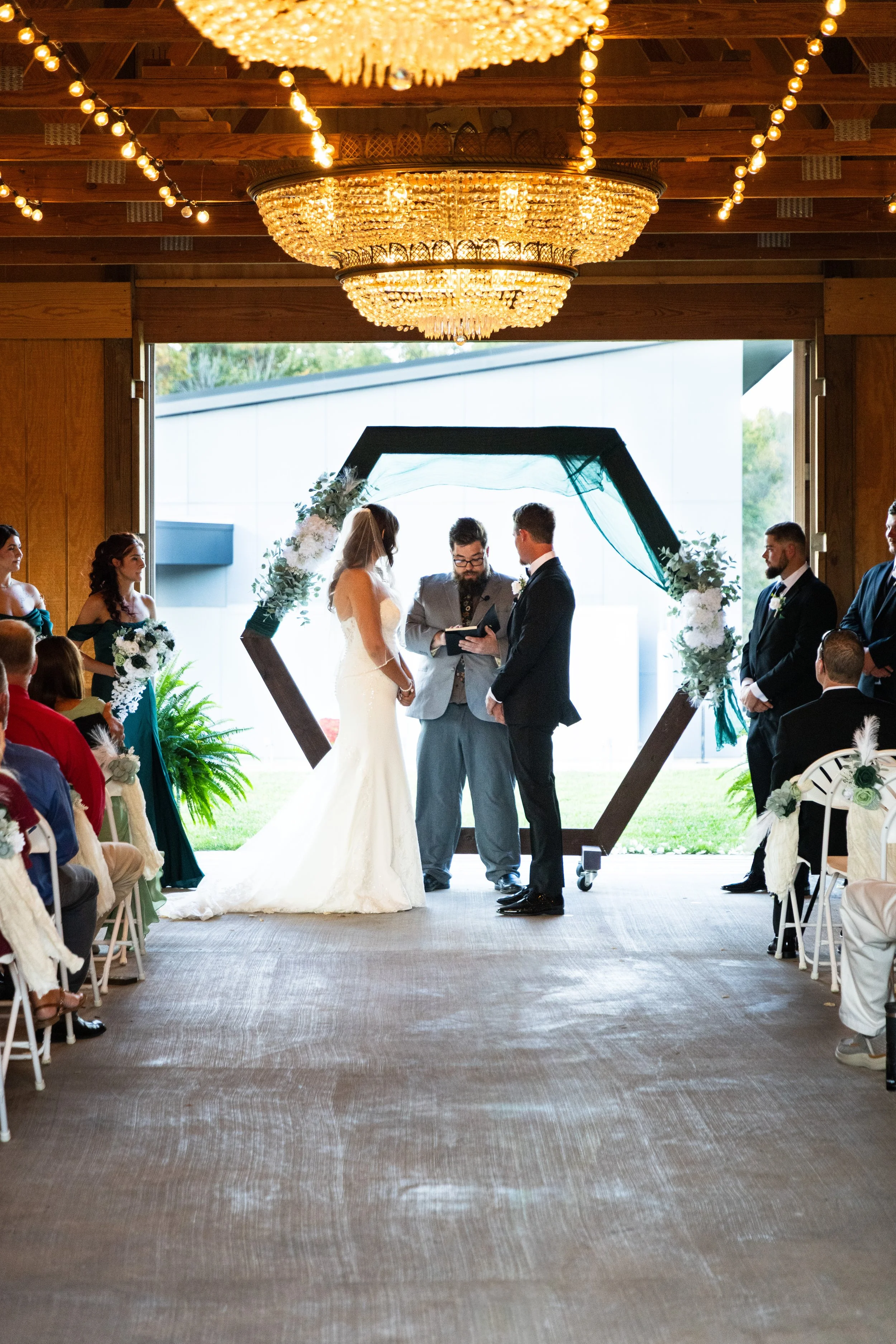 A wedding ceremony taking place indoors with a couple standing before an officiant under a hexagonal floral and fabric archway. The bride is in a white gown and veil, and the groom is in a black suit. Guests are seated on either side, watching the ceremony. The room has wooden walls, a large hanging chandelier, and string lights.