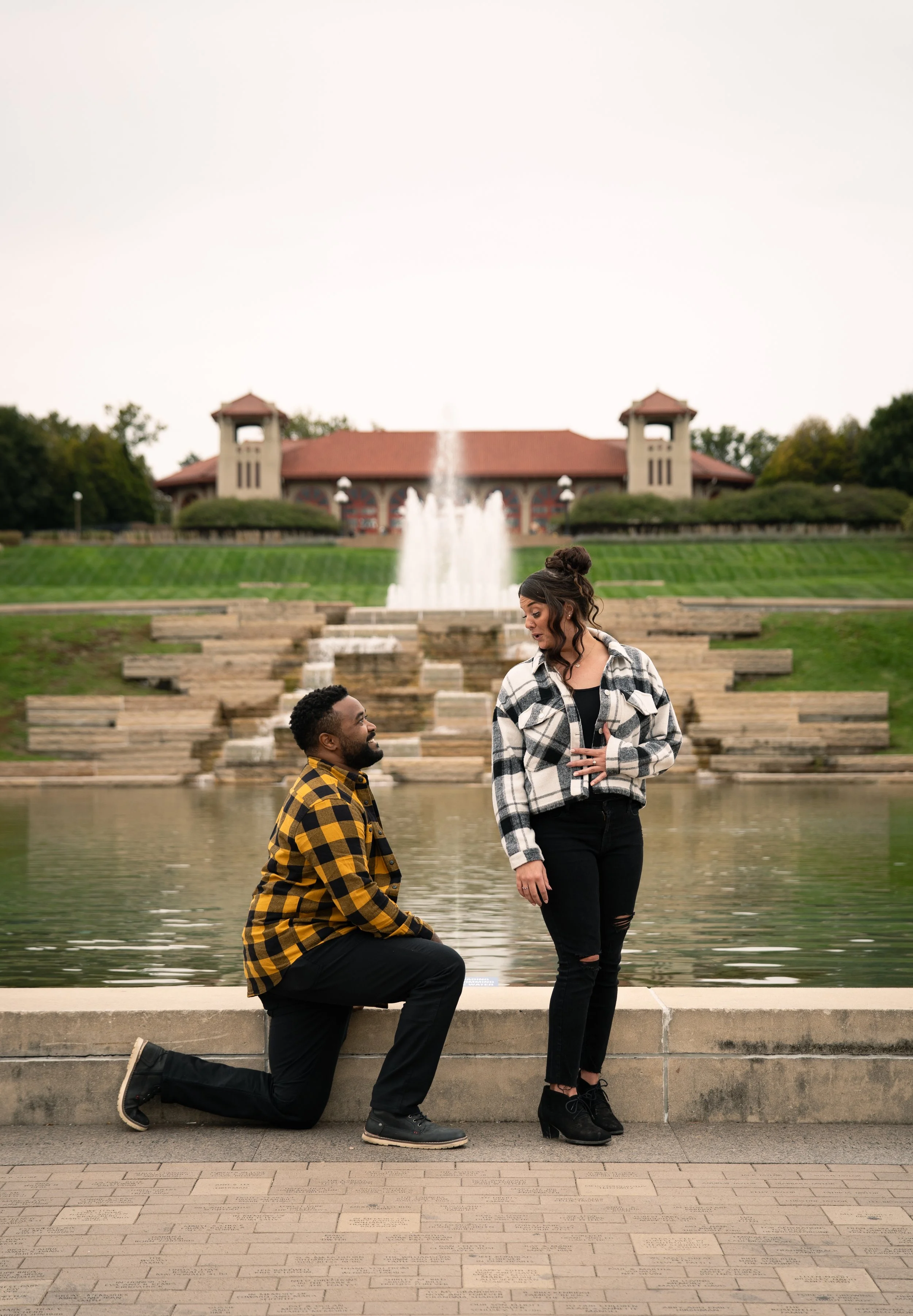 A man proposing marriage to a woman by a fountain in a park with a building and fountain in the background.