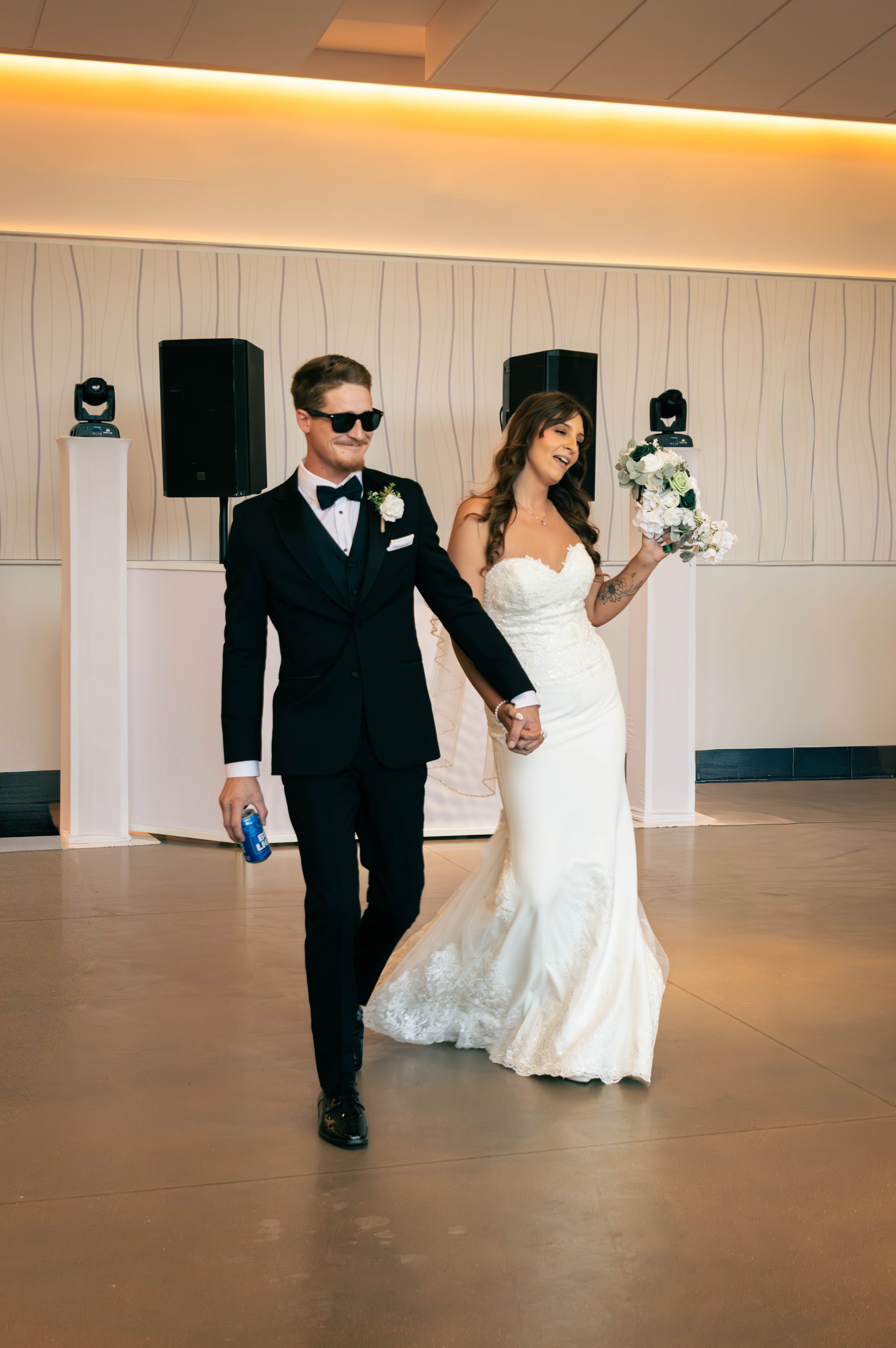 A newlywed couple holding hands and walking at their wedding reception in a modern room, with the bride in a white lace wedding dress holding a bouquet of white flowers and the groom in a black tuxedo with sunglasses, a bow tie, and a boutonniere.